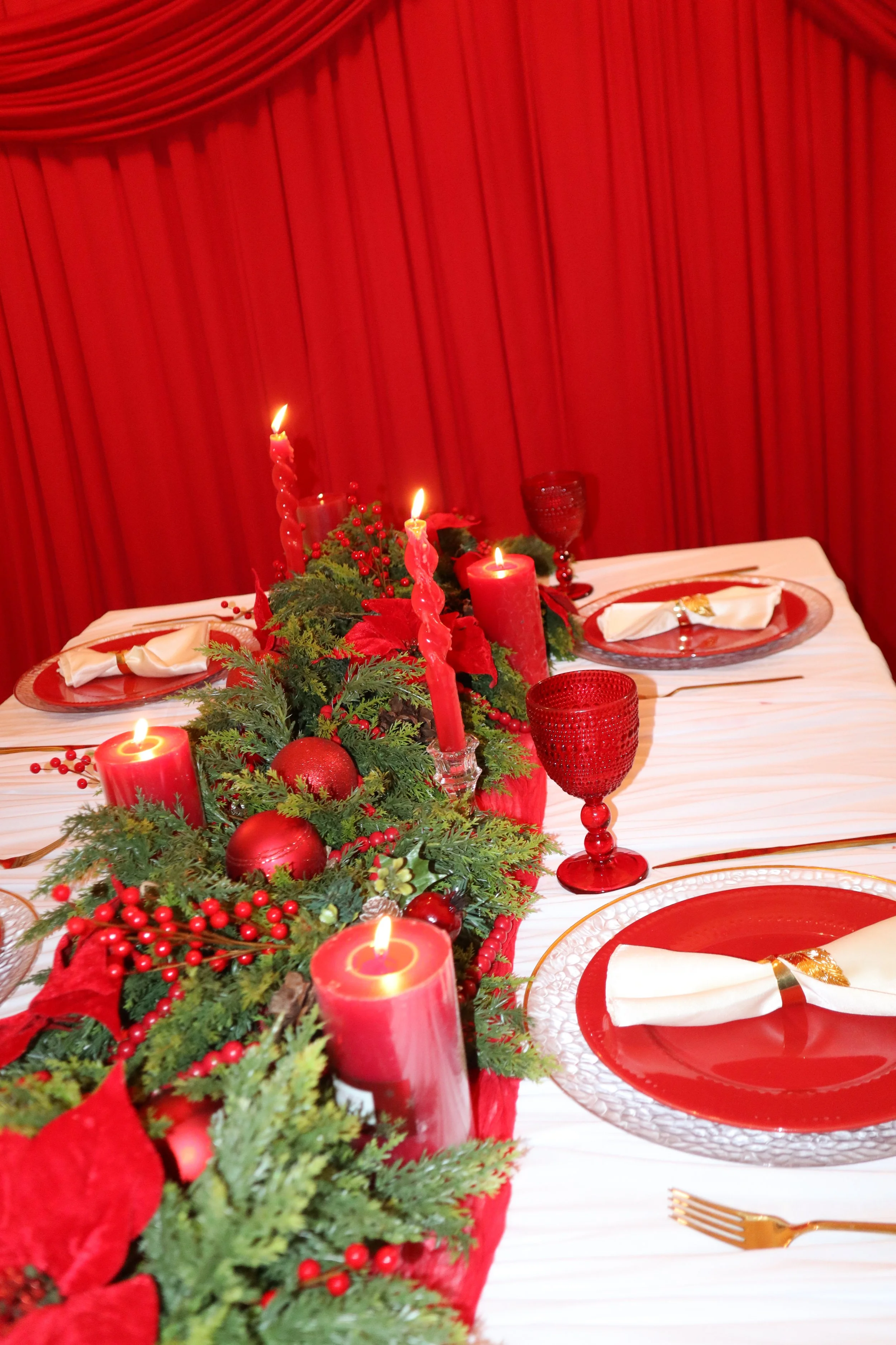 A festive Christmas dinner table decorated with red candles, red and gold tableware, and green foliage with red berries and ornaments. A red curtain hangs in the background.