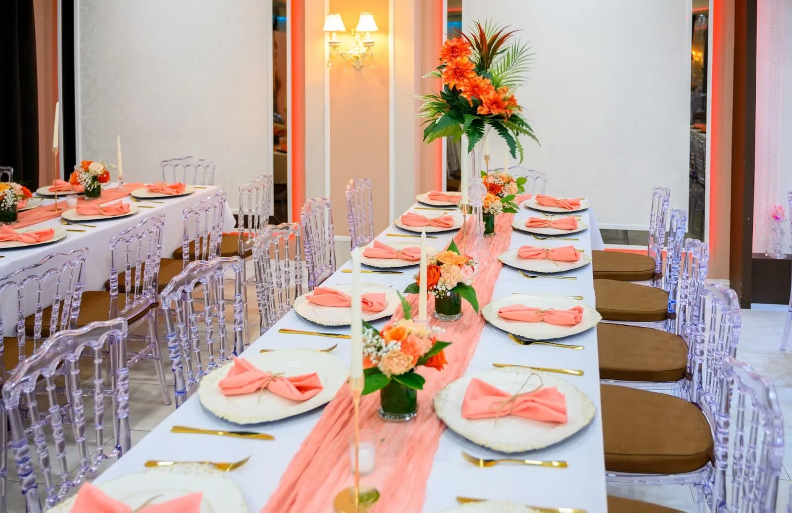 A decorated banquet table for a celebration with floral centerpieces, pink napkins, and candles, in a room with elegant lighting and white walls.