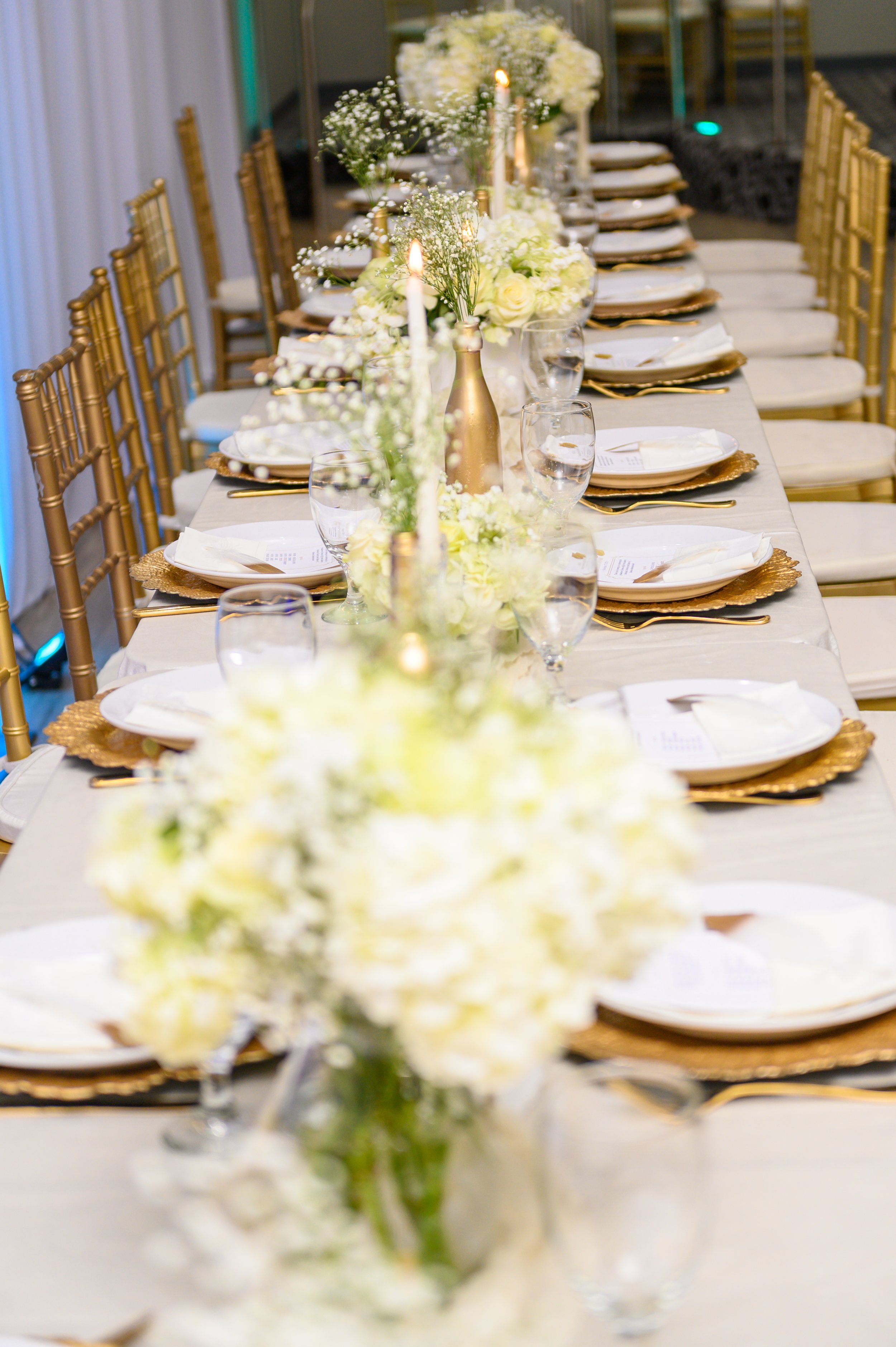 A long table set for a formal event, decorated with floral centerpieces, candles, gold chargers, white napkins, and glassware, with gold chairs surrounding it.