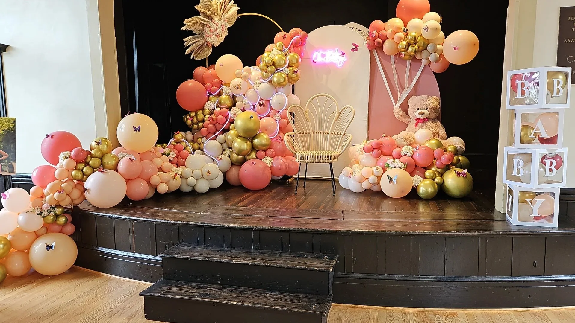 Decorative setup with pink, white, and gold balloons, a rattan chair, a teddy bear holding balloons, flower arrangements, and a neon sign reading 'Angel' on a small stage.