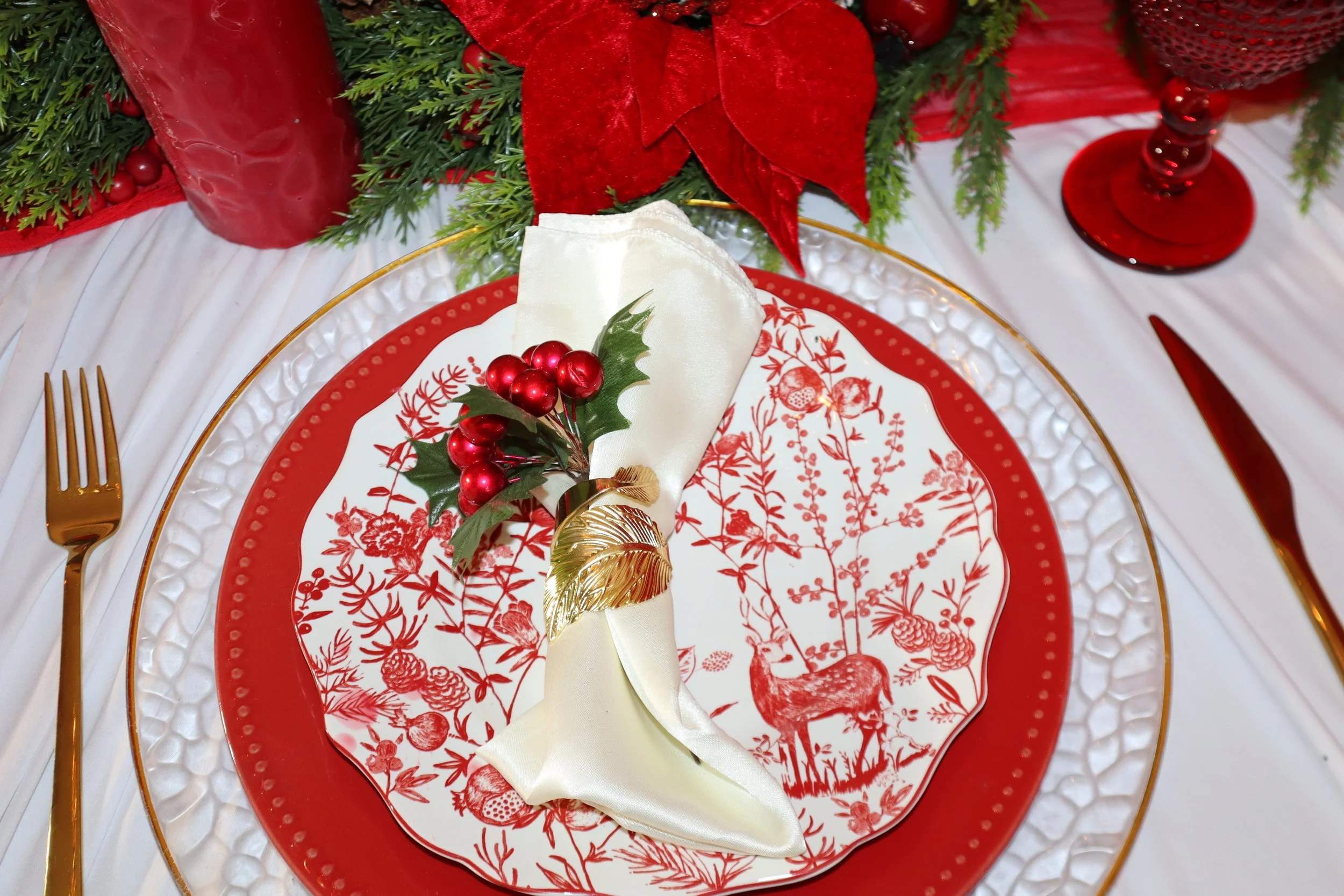 A Christmas-themed table setting featuring a red and white patterned plate, a cream-colored napkin, and festive decorations including red berries, holly leaves, and poinsettia flowers. Gold utensils are placed beside the plate, with greenery and red 