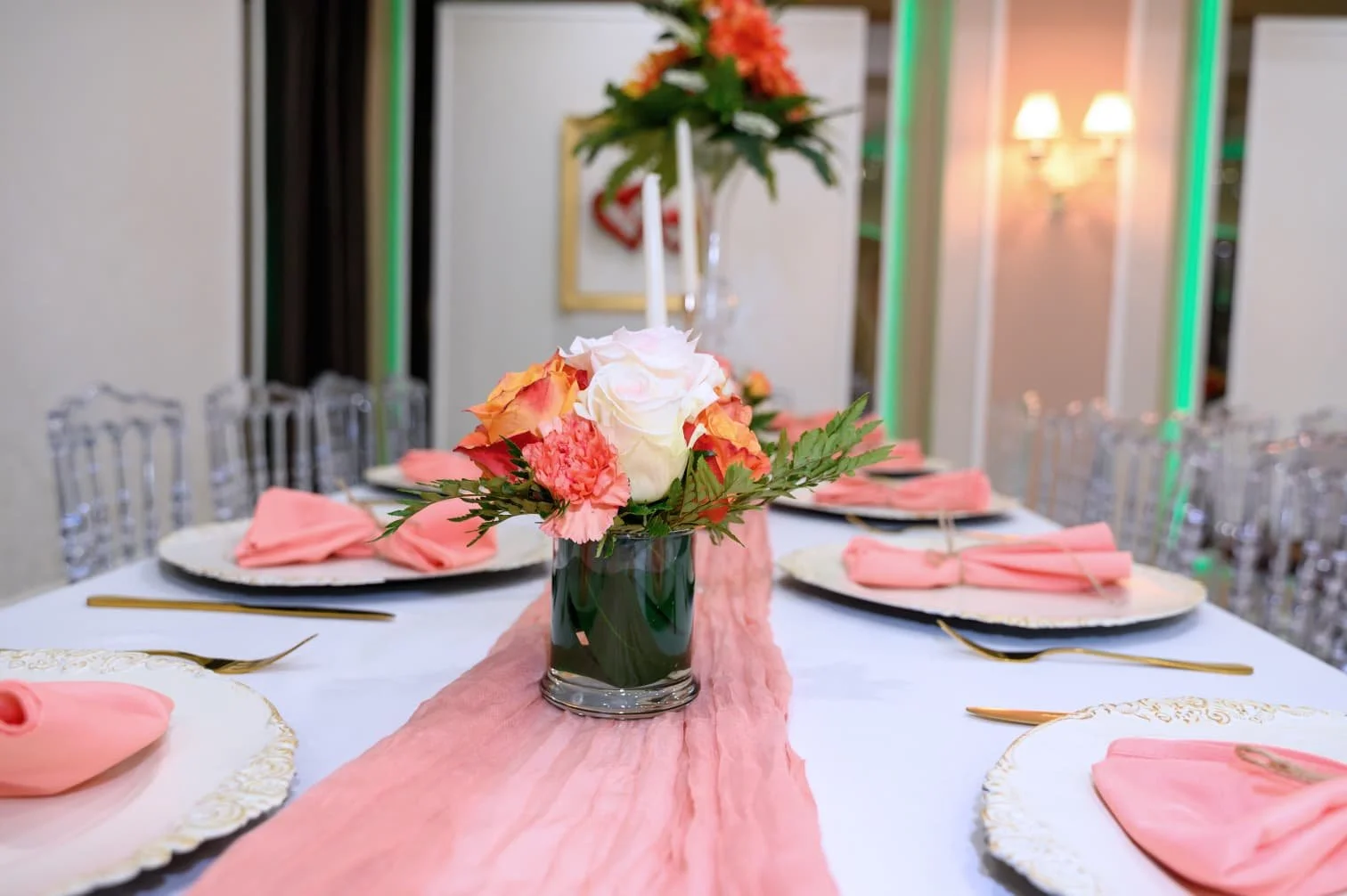 A dining table set with a pink table runner, pink napkins, and gold utensils, with a floral centerpiece including white roses and pink flowers in a glass jar, in an elegantly decorated room.