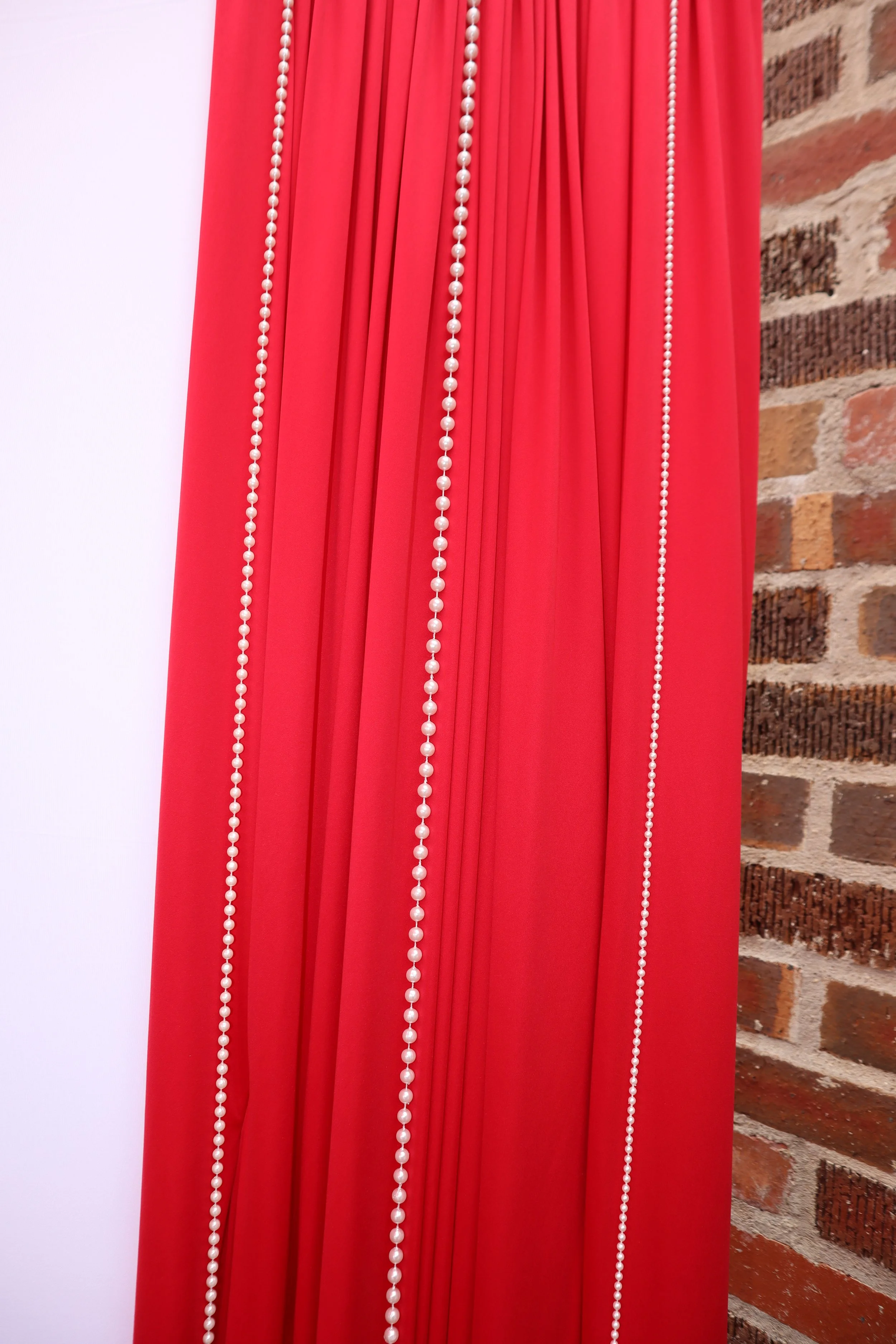 Red curtains with white pearl-like beads hanging vertically, against a background of a brick wall on the right and a plain white wall on the left.