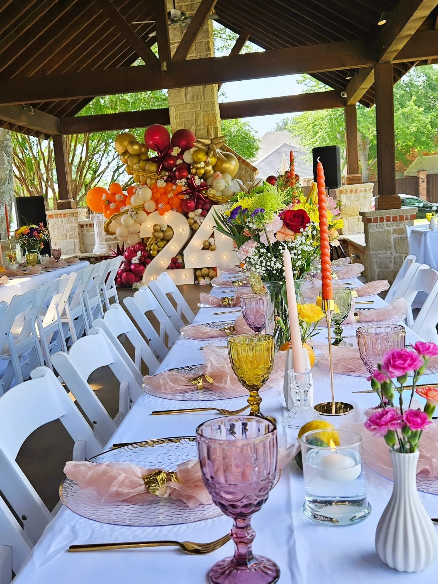 A decorated outdoor banquet table with pink and purple glasses, floral arrangements, lit candles, and a large '2024' balloon centerpiece, under a wooden pavilion surrounded by trees.