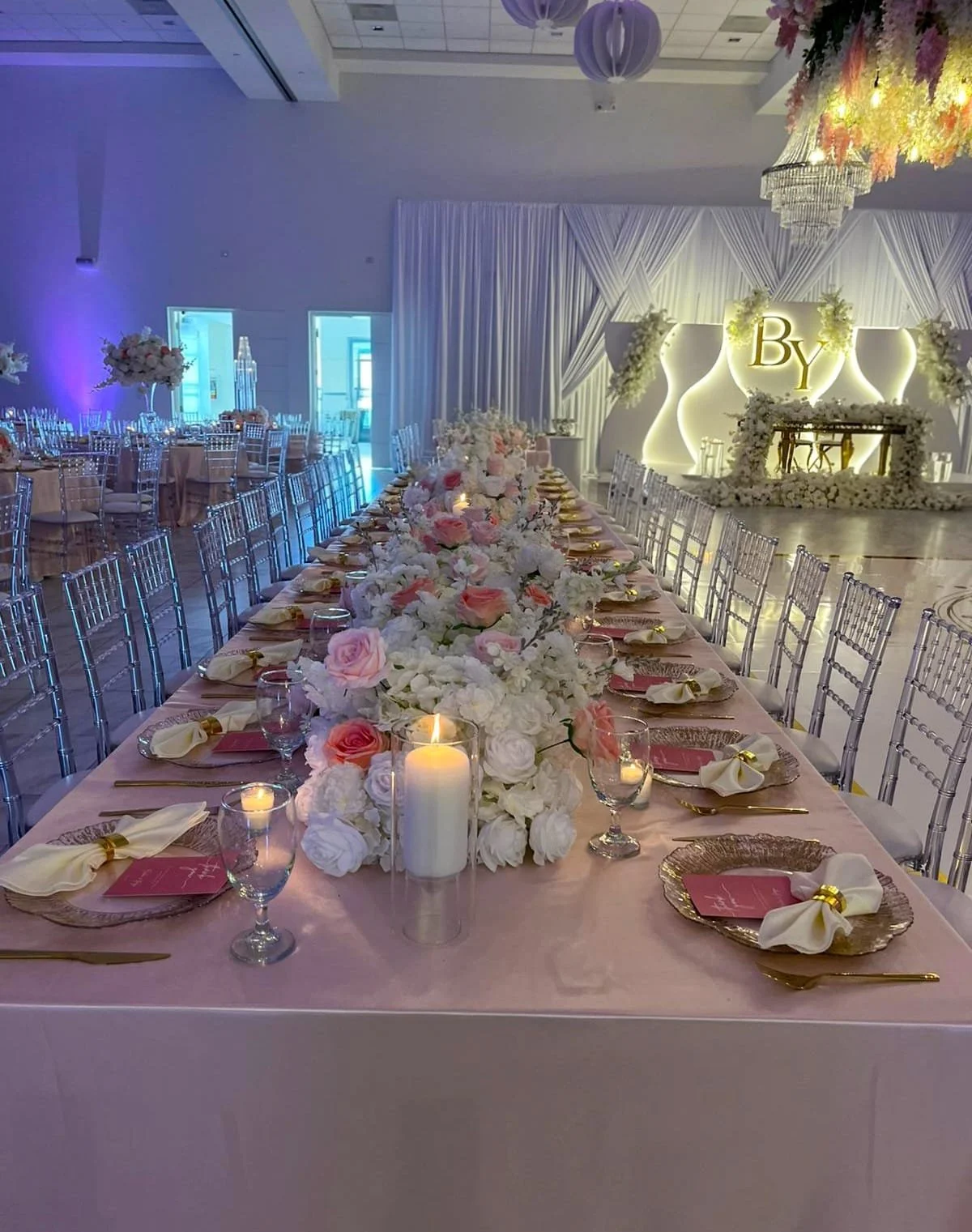 A banquet table decorated with pink and white flowers, candles, and gold-edged plates, set for a wedding reception with a decorated stage in the background featuring the letter 'B'.