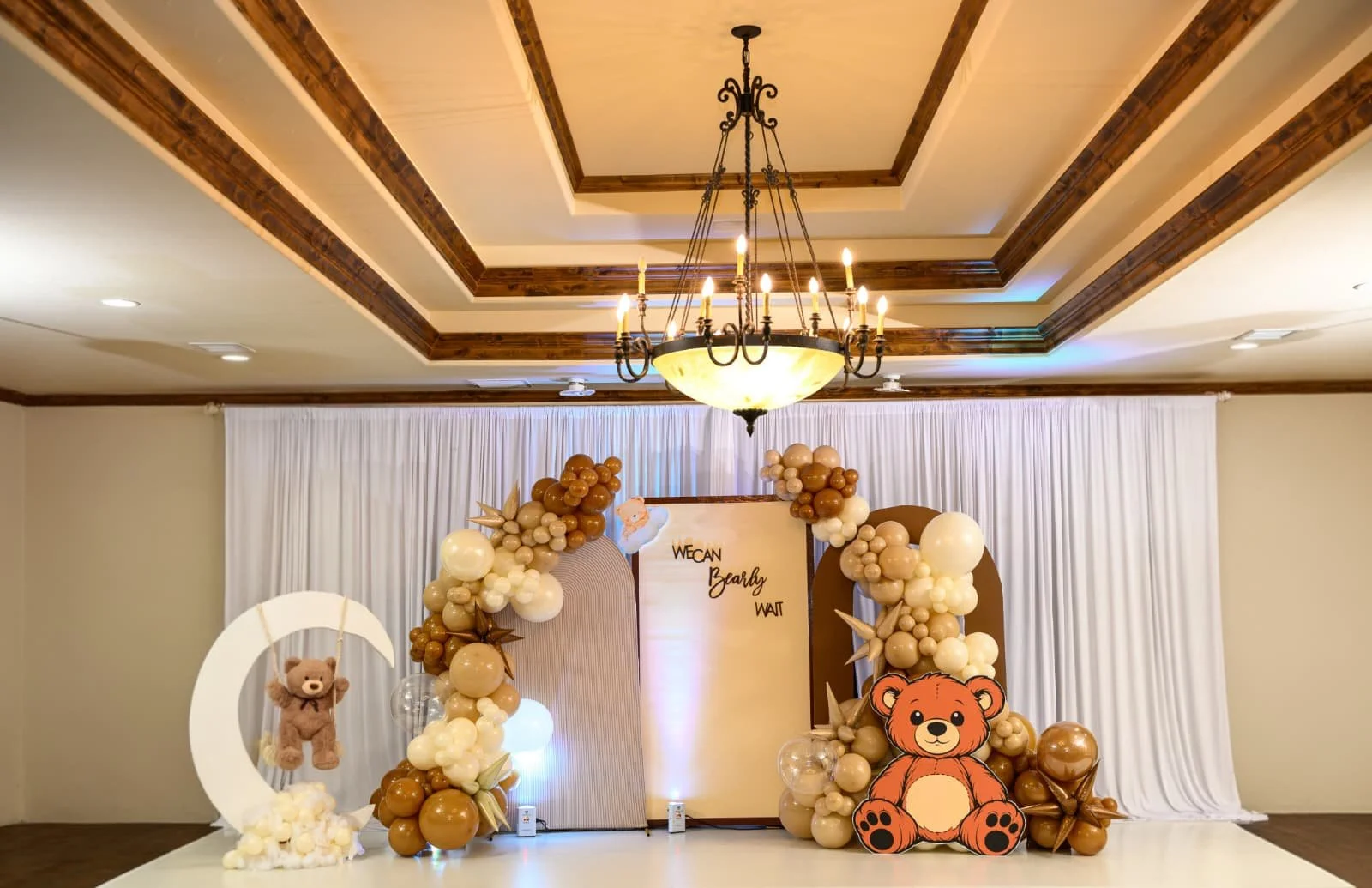 Decorations for a baby shower or celebration featuring a balloon arch with teddy bear and moon cutouts, a white curtain backdrop, and a large teddy bear cutout at the front of the stage, with a chandelier hanging from the ceiling.