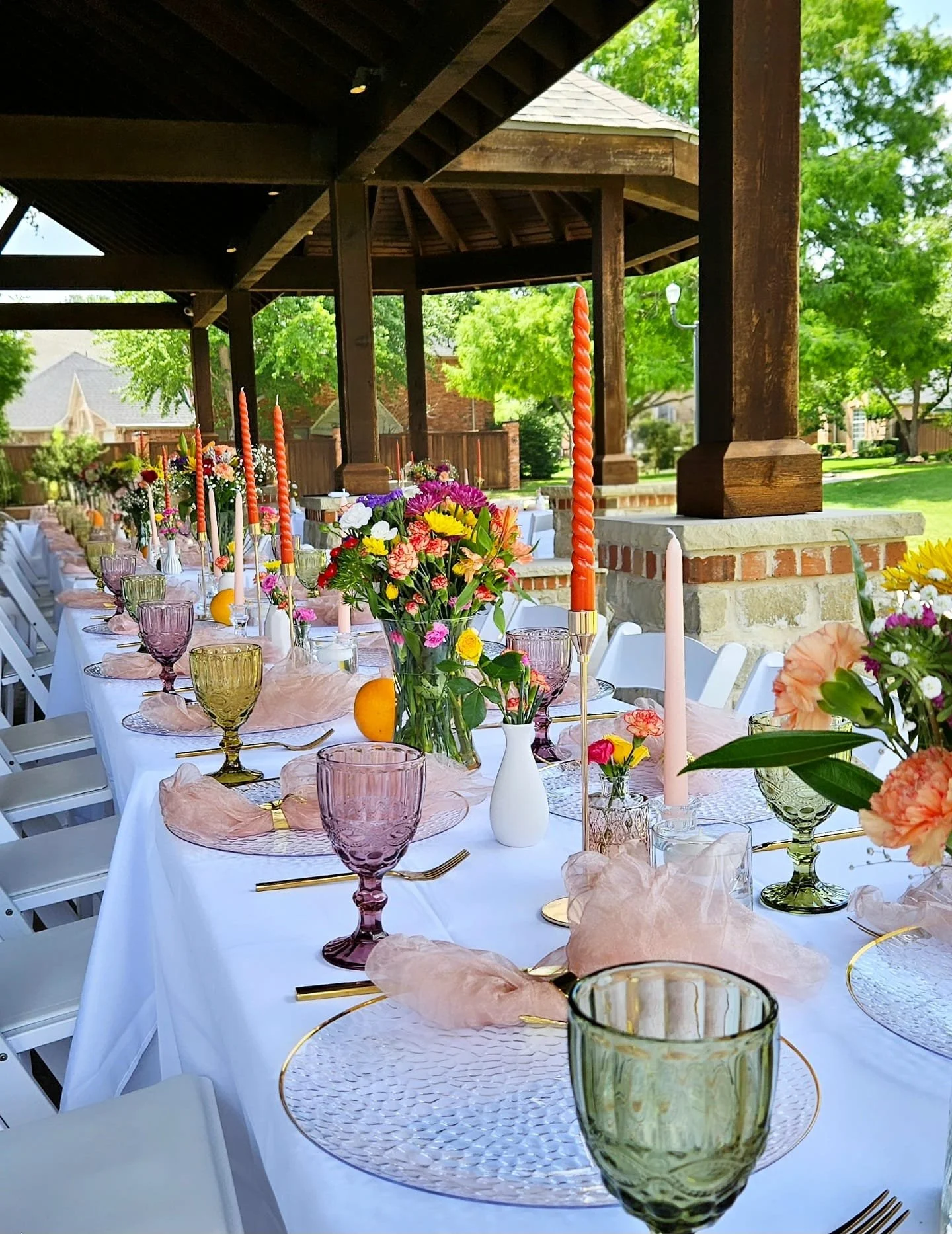 Decorated outdoor dining table with pink, purple, yellow, and white flowers, colorful candles, glassware, and gold utensils, under a wooden pavilion with green trees in the background.
