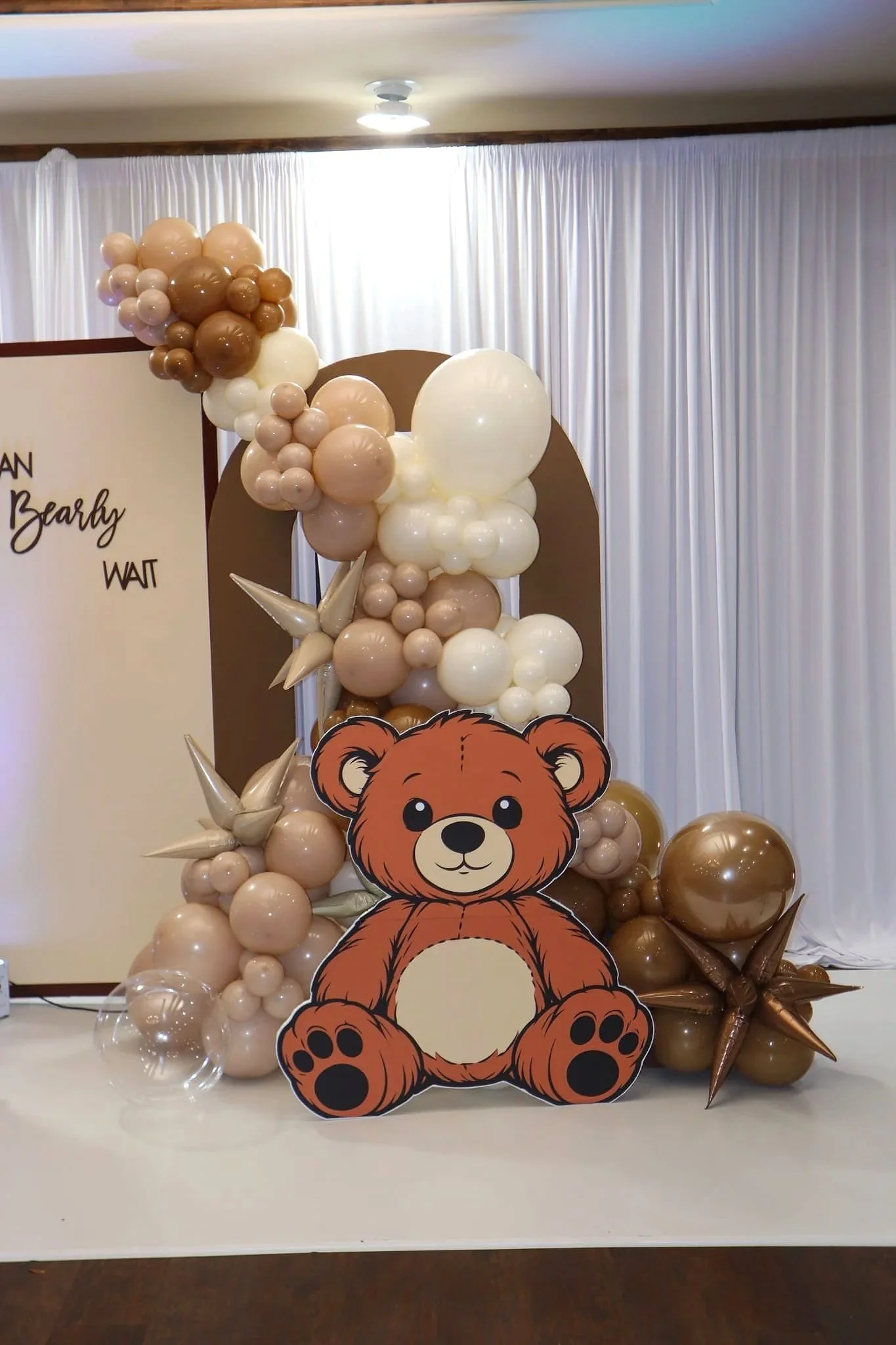 Decorative balloon arrangement with shades of brown, beige, and white balloons, featuring a cute teddy bear cutout at the front, set against a background of white curtains and a whiteboard with partial writing.