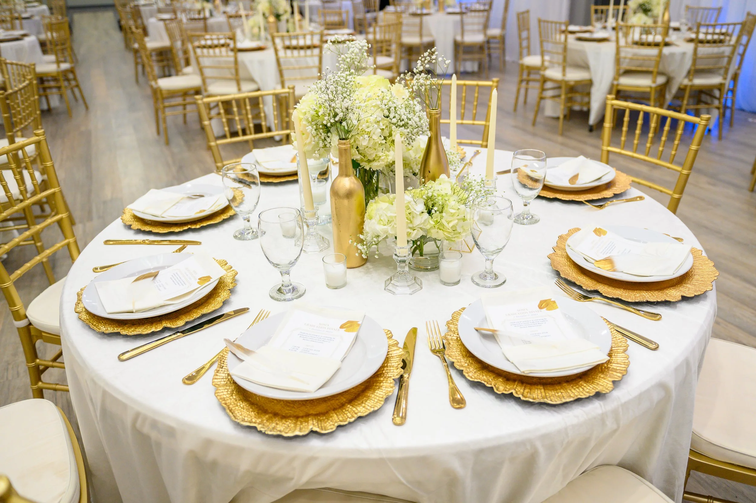 A round banquet table decorated with gold placemats, white plates, gold utensils, clear wine glasses, and white napkins. The centerpiece consists of white flowers in gold vases and white candles. The table is set in a formal event space with gold chairs and additional tables in the background.