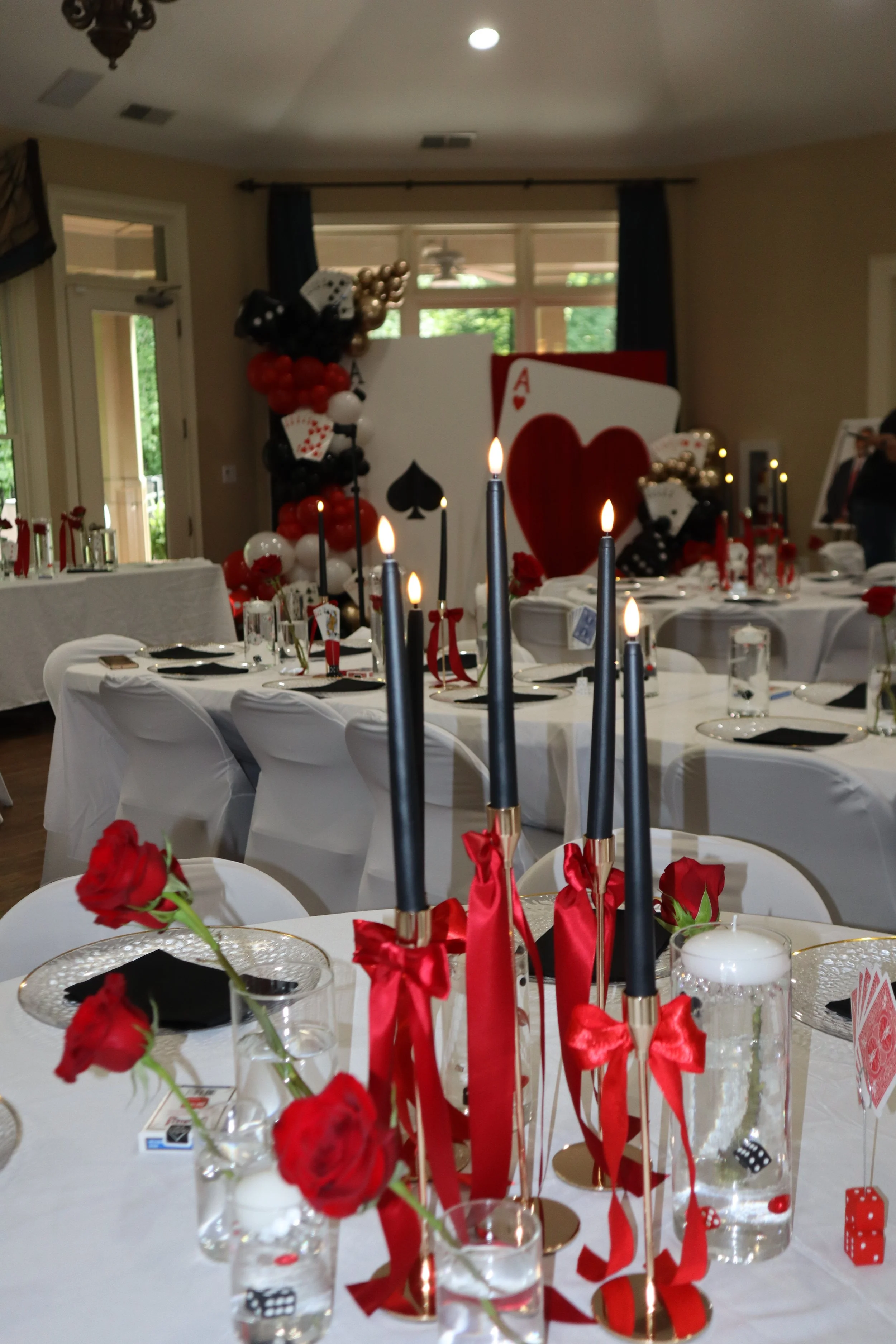 Party table decorated with red flowers, black and red ribbons, candles with red ribbons, game dice, and a large playing card with a red heart symbol, against a background with balloon decorations and a large love-themed card display.