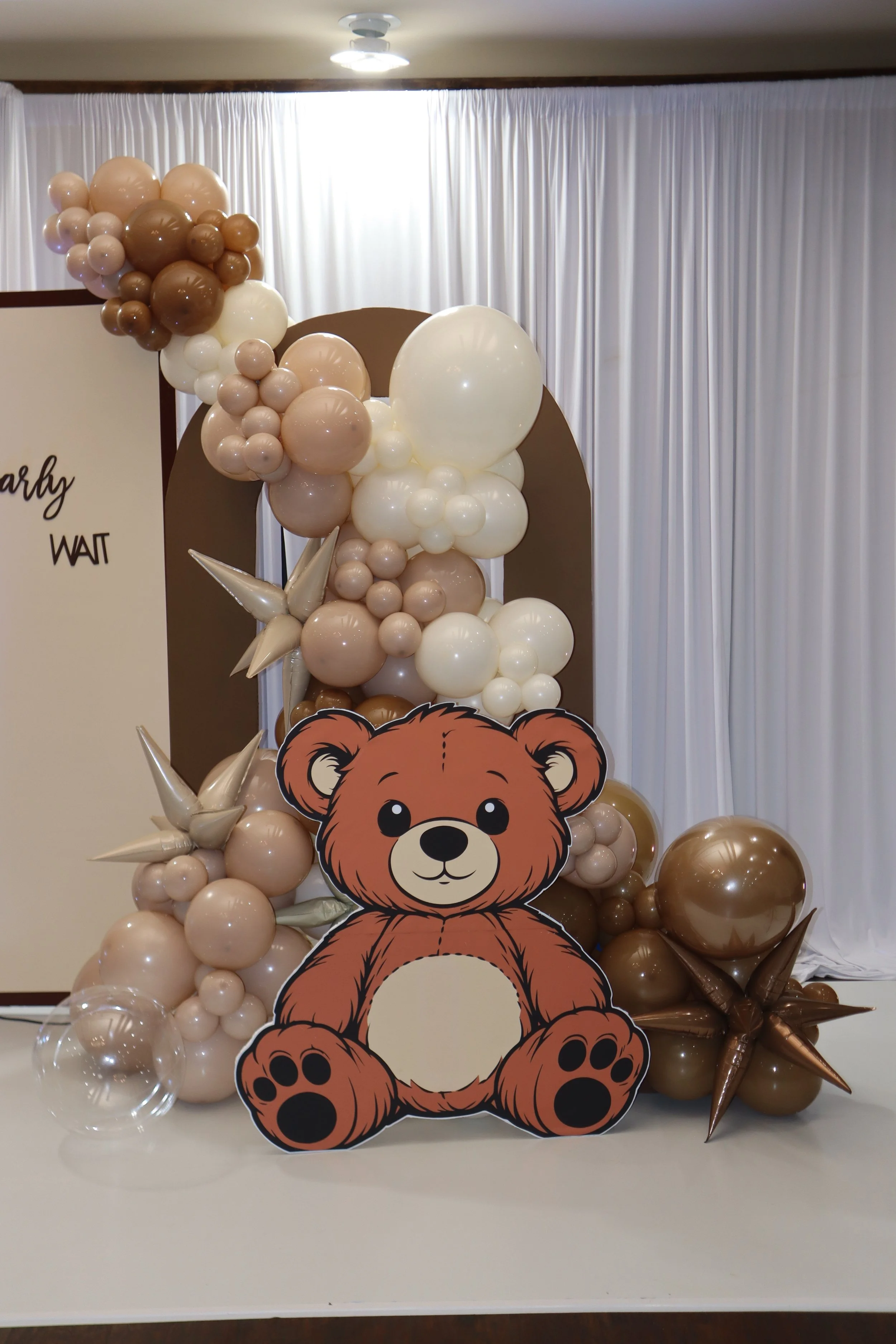 Decorative bear cutout surrounded by tan, white, and brown balloons with white curtain backdrop.