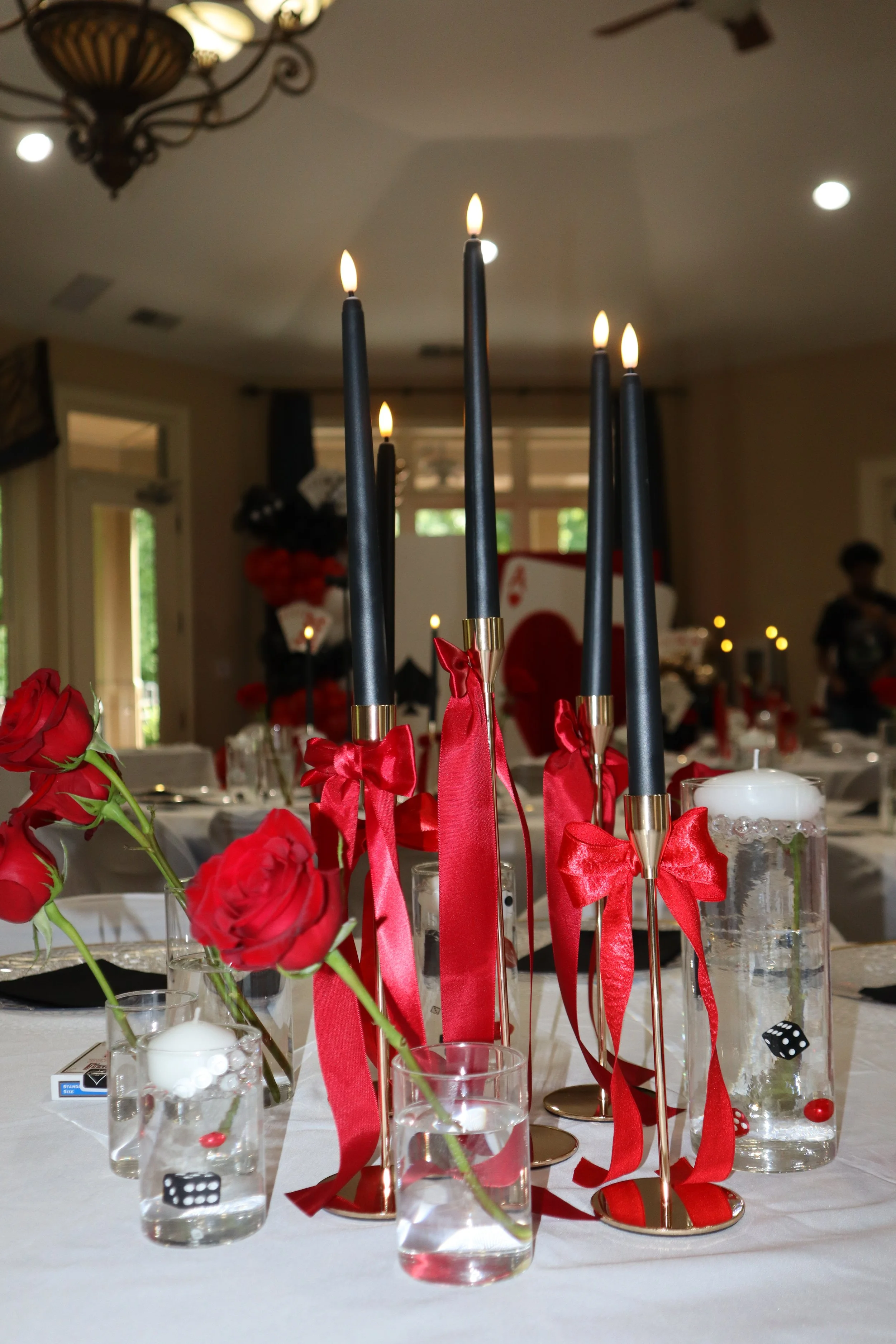 Table decorated with black candles, red ribbons, red roses, and small glass vases with white candles inside. The background shows a dimly lit room with a chandelier and some blurred figures and decorations, suggesting a celebration or party setting.