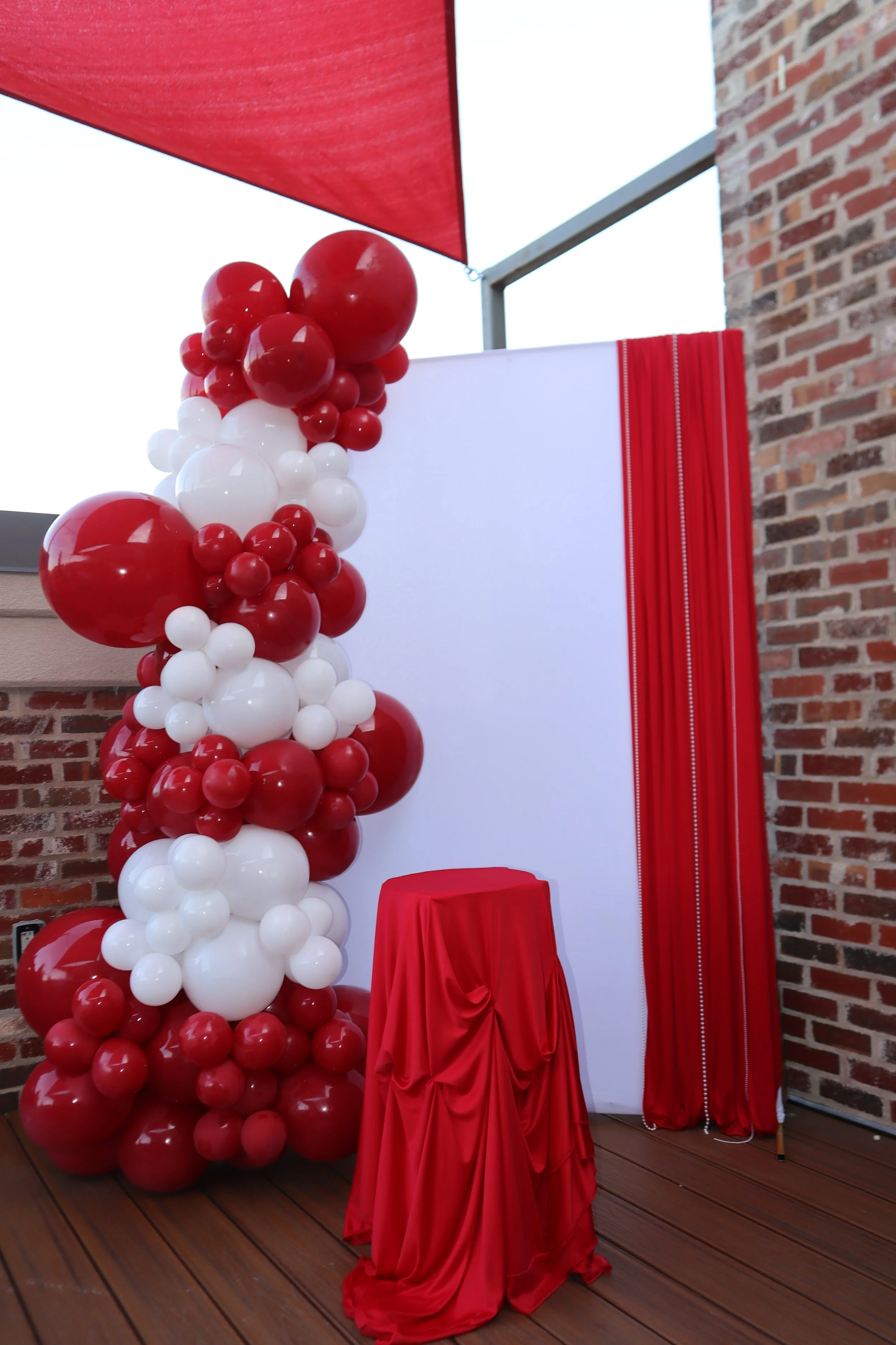 Decorative setup with a large column of red and white balloons, a red-draped table, and red and white fabric backdrop on a wooden floor patio with brick wall and metal railing.