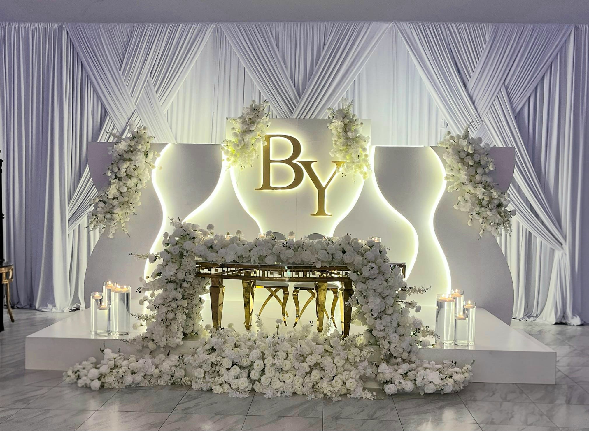 Elegant wedding or event backdrop with white drapery, illuminated initials 'B' and 'Y' in gold, and abundant white floral arrangements with candles.