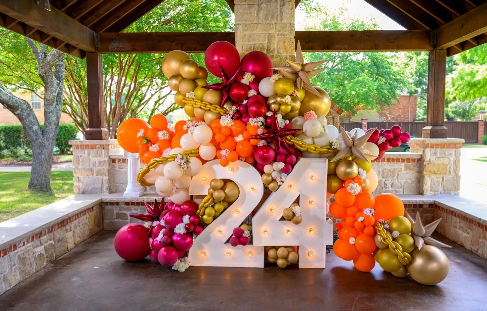 A decorative balloon arrangement in the shape of the number 24, featuring large balloons in red, white, gold, and orange, accented with flower and star-shaped balloons, set against a pavilion outdoor with trees and brickwork in the background.