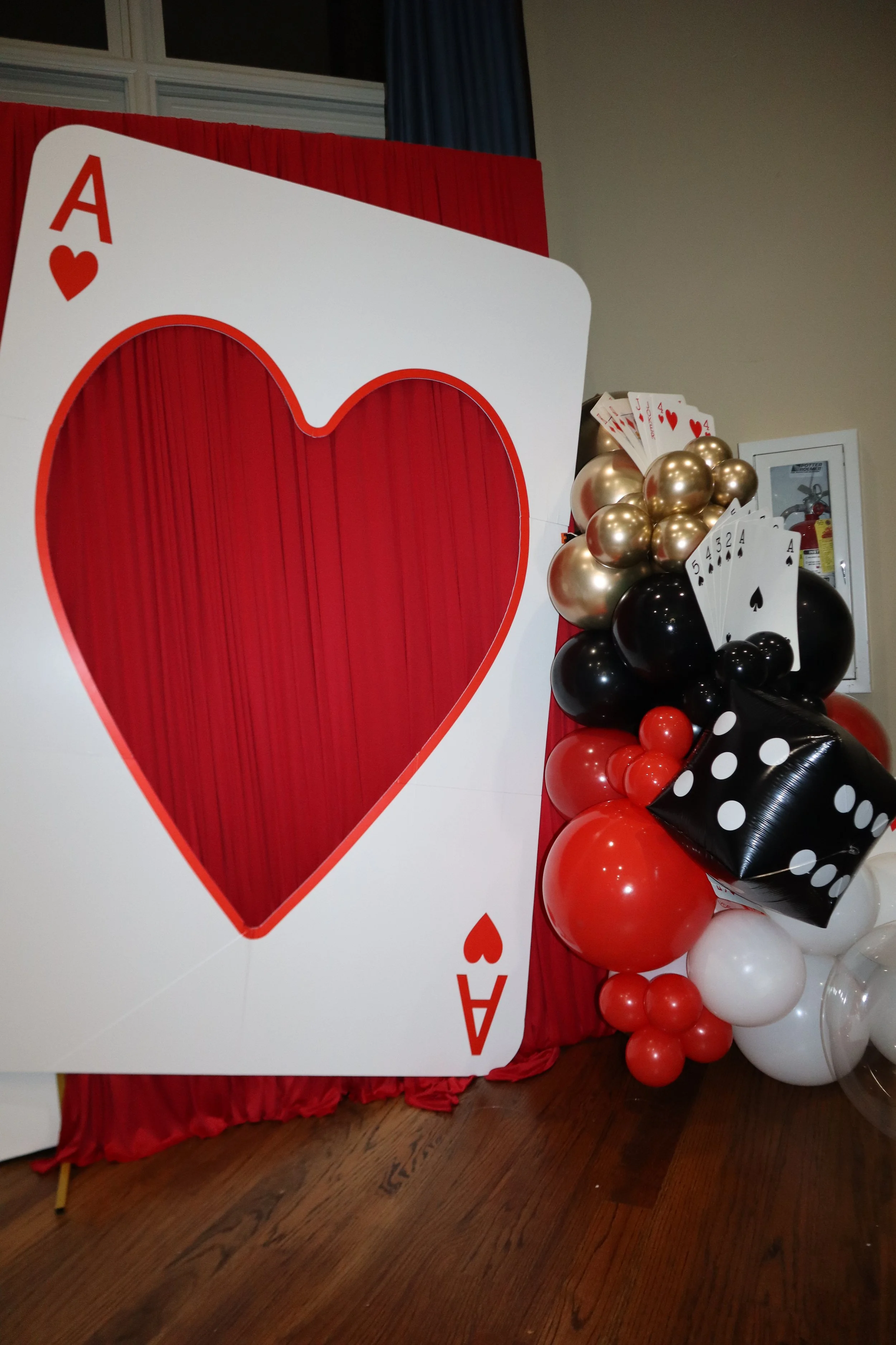 A large cardboard cutout of an Ace of hearts card with a heart shape cutout in the center, decorated with black, red, and white balloons arranged in a cluster, on a red table with a red and blue curtain in the background.
