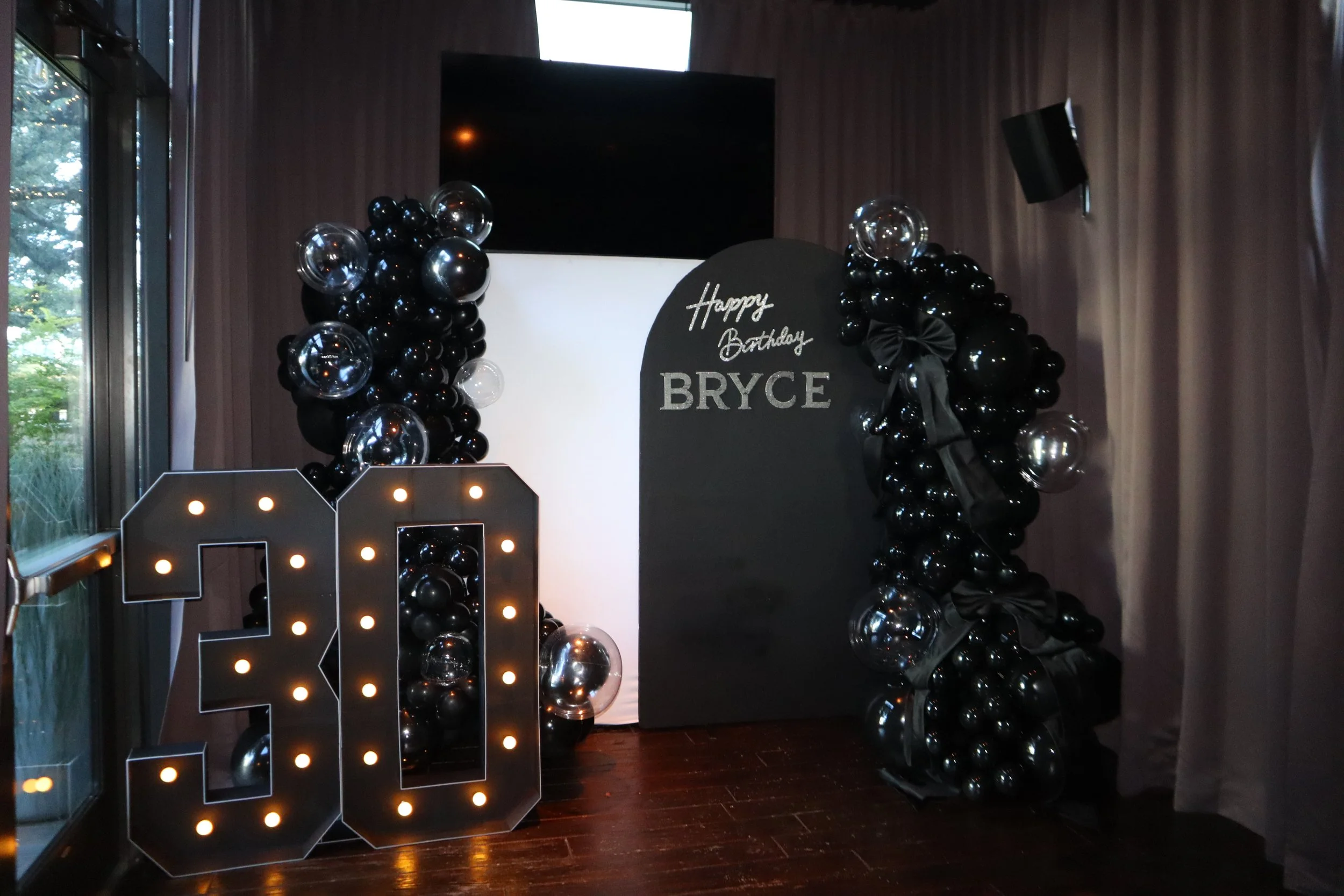 Black balloon arch with transparent balloons on each side surrounding a black sign that says "Happy Birthday Bryce" in silver lettering, with black ribbon decorations, on a dark wood floor near a window.