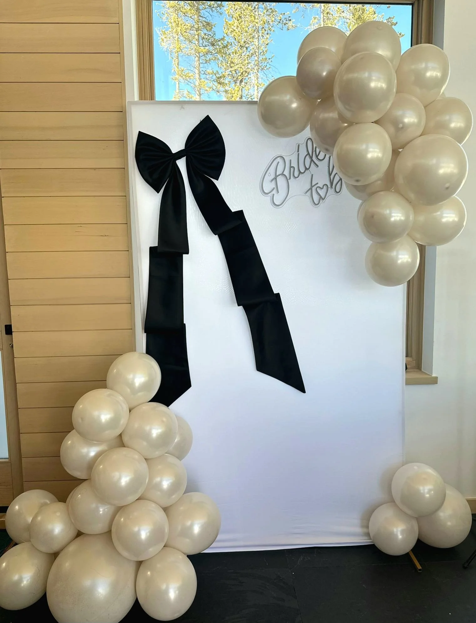 Decorative photo with white balloons forming two columns, a black ribbon bow on a textured white backdrop, and the words 'Bride to Be' written on the backdrop. A window showing trees and blue sky is in the background.