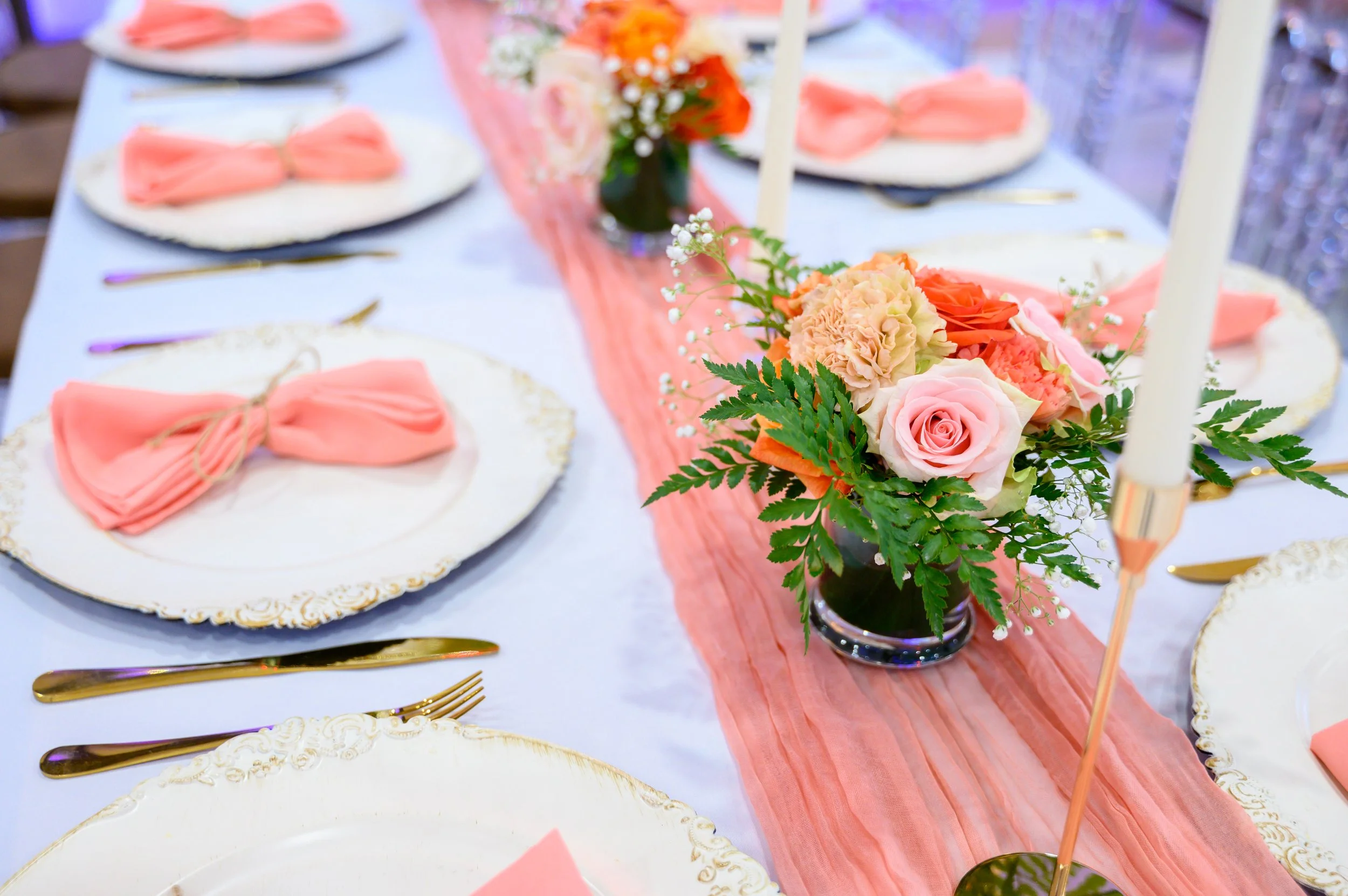 A decorated dining table with pink and white floral centerpiece, white plates with pink napkins, and gold cutlery, set for a special occasion.