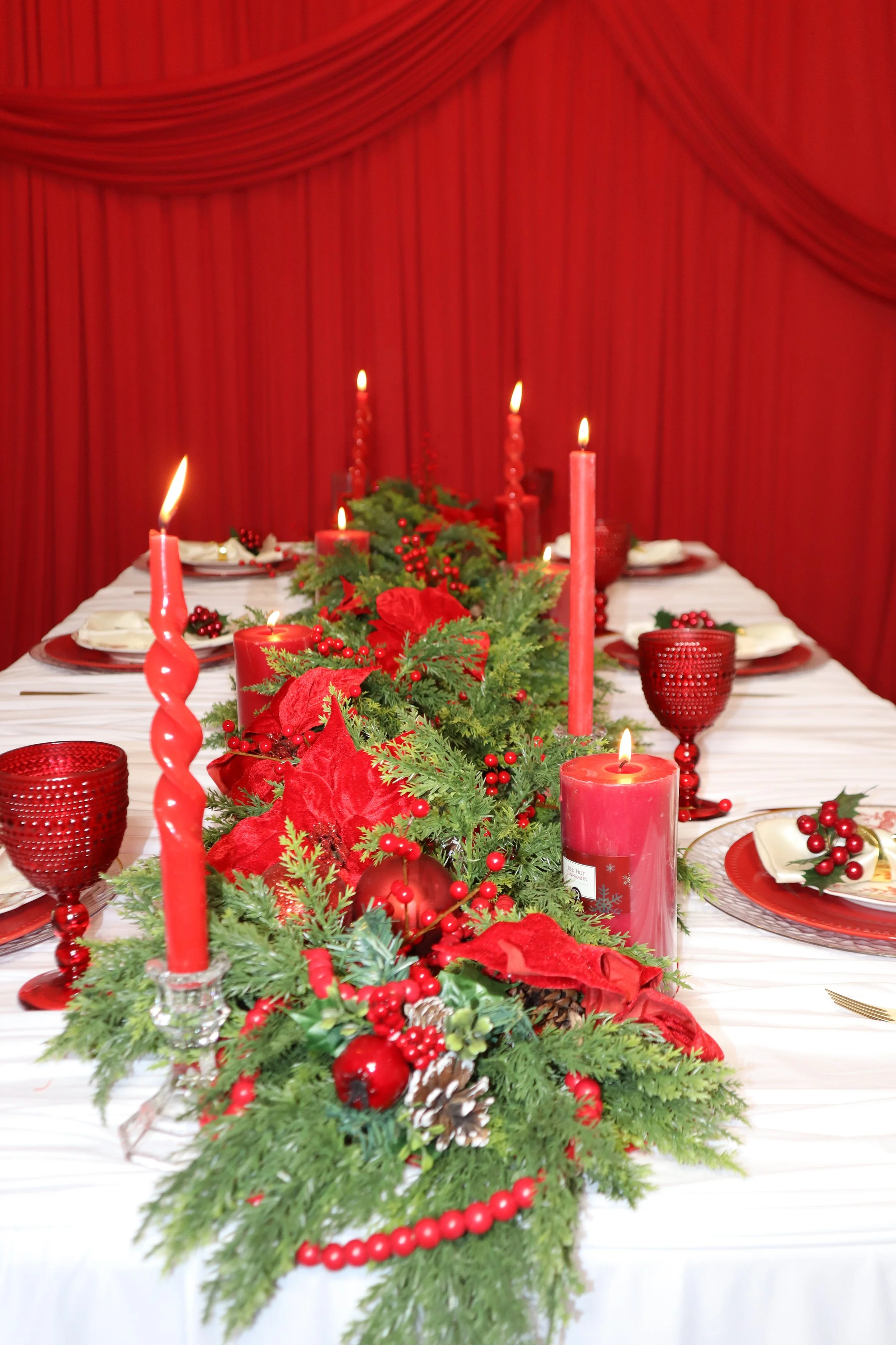 Christmas table centerpiece with red candles, greenery, and red holiday decorations on a white table with red glassware and red napkins, against a red curtain backdrop.