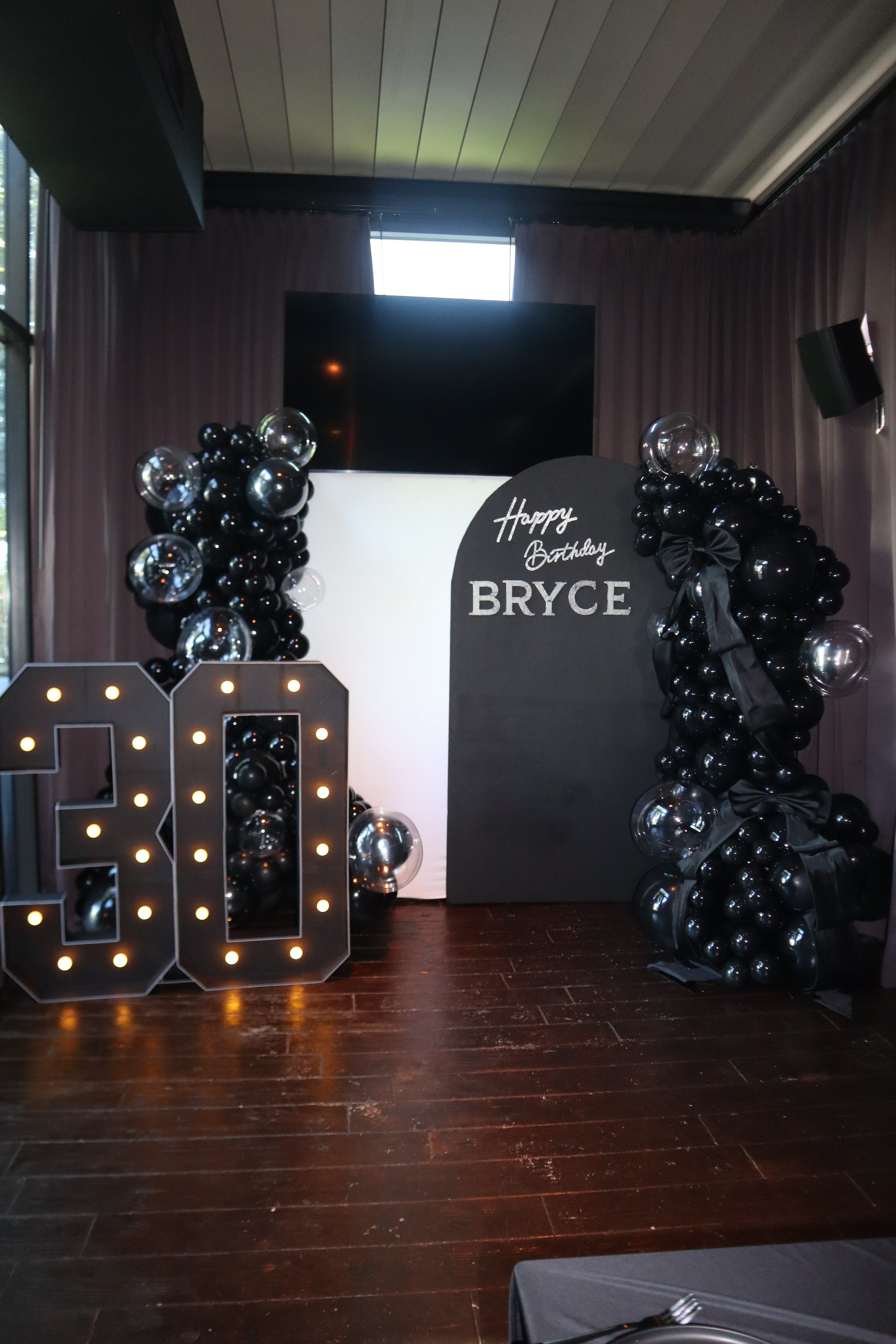 A decorated birthday celebration setup with large illuminated letter numbers, balloon arrangements in black and silver, and a black chalkboard style sign that reads 'Happy Birthday BRYCE'.