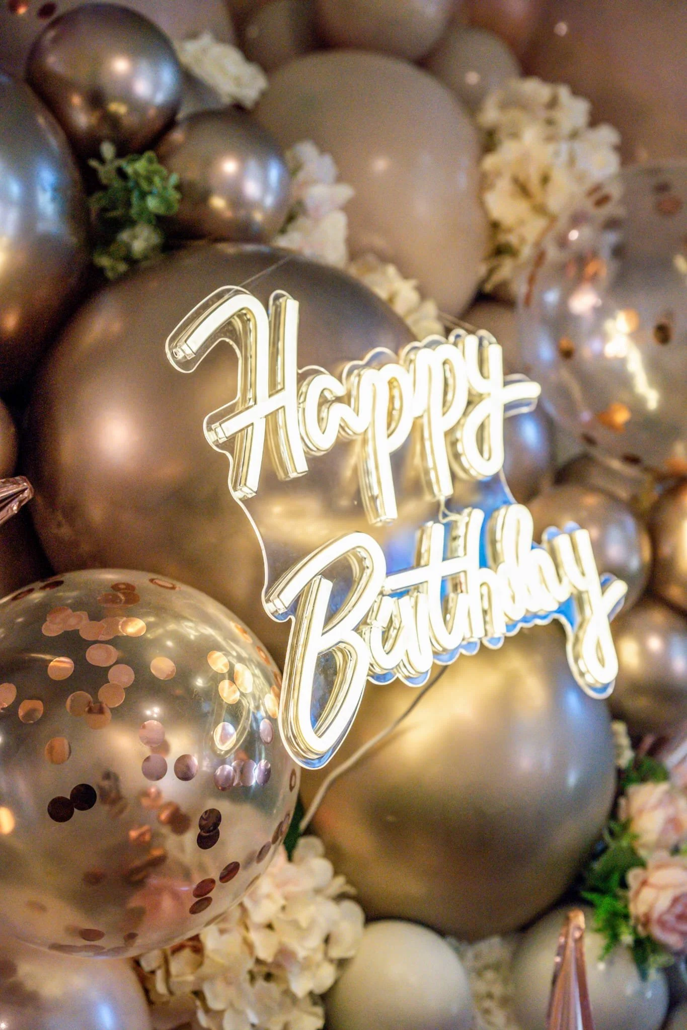 A close-up of a birthday decoration display featuring a shiny metallic balloon, a transparent balloon with pink and gold confetti inside, white popcorn, green leaves, and a glowing neon sign that reads 'Happy Birthday'.