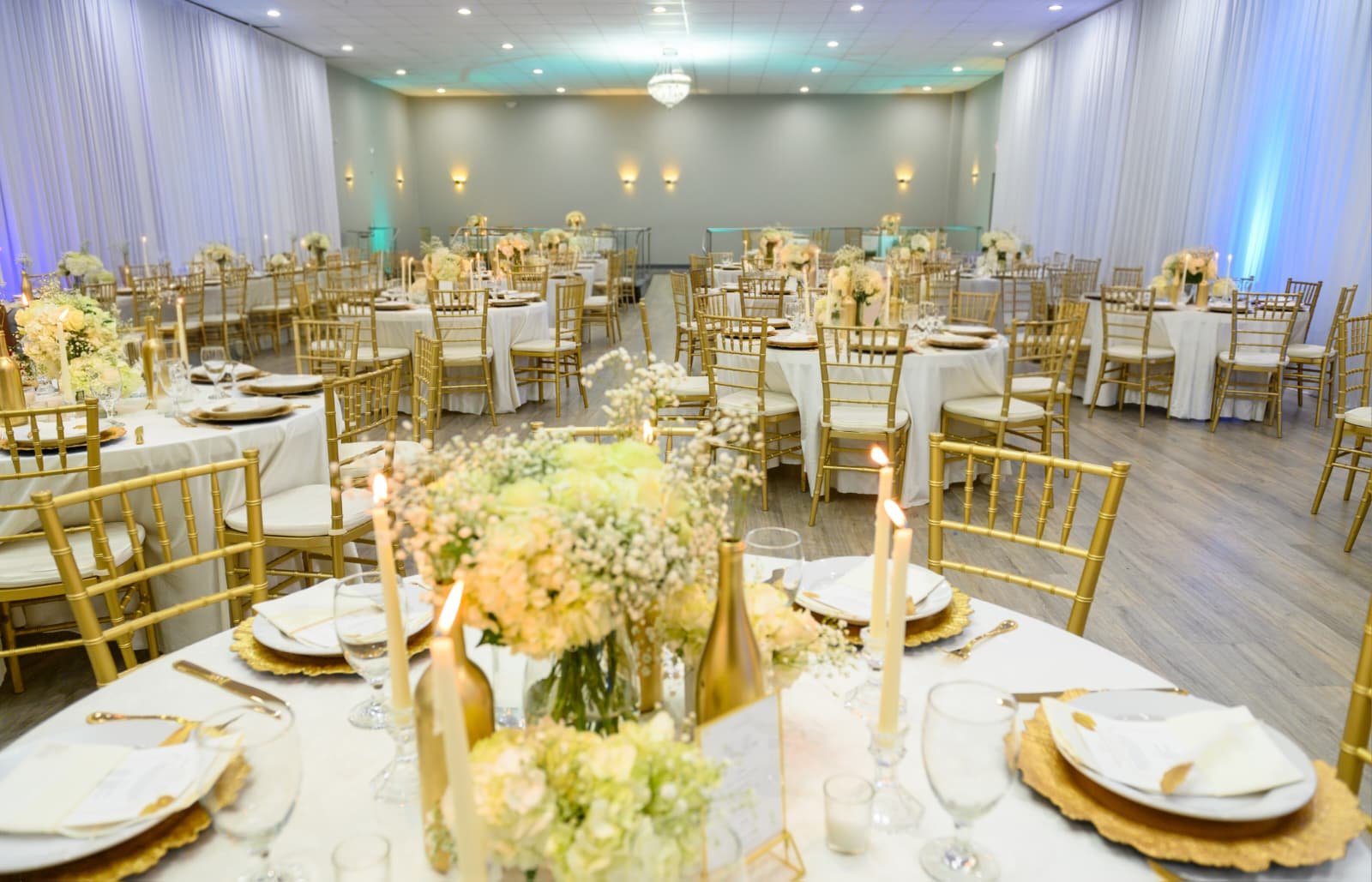 A banquet hall decorated for a formal event with round tables with white tablecloths, gold chairs, floral centerpieces, candles, and place settings.