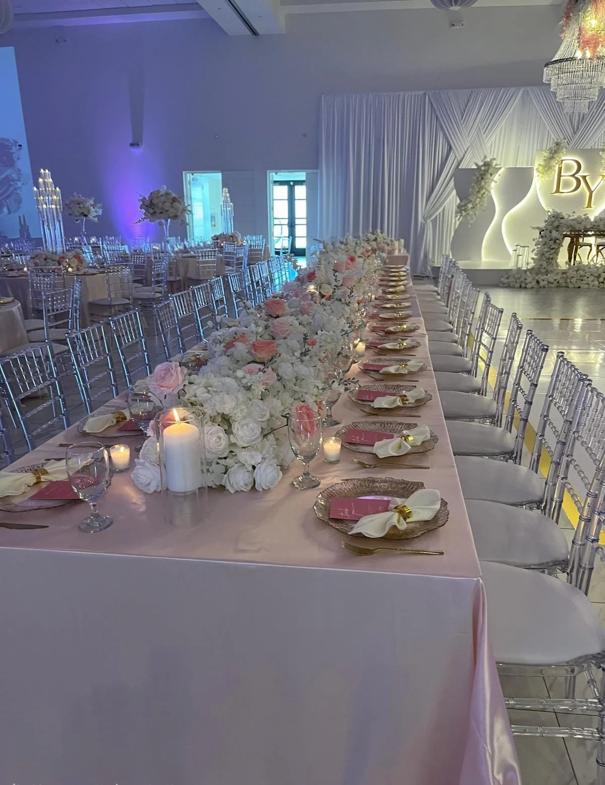 Formal event setup with a long table decorated with white and pink flowers, candles, and gold accents, with chairs arranged on both sides, and a stage in the background with a white backdrop and floral decorations.