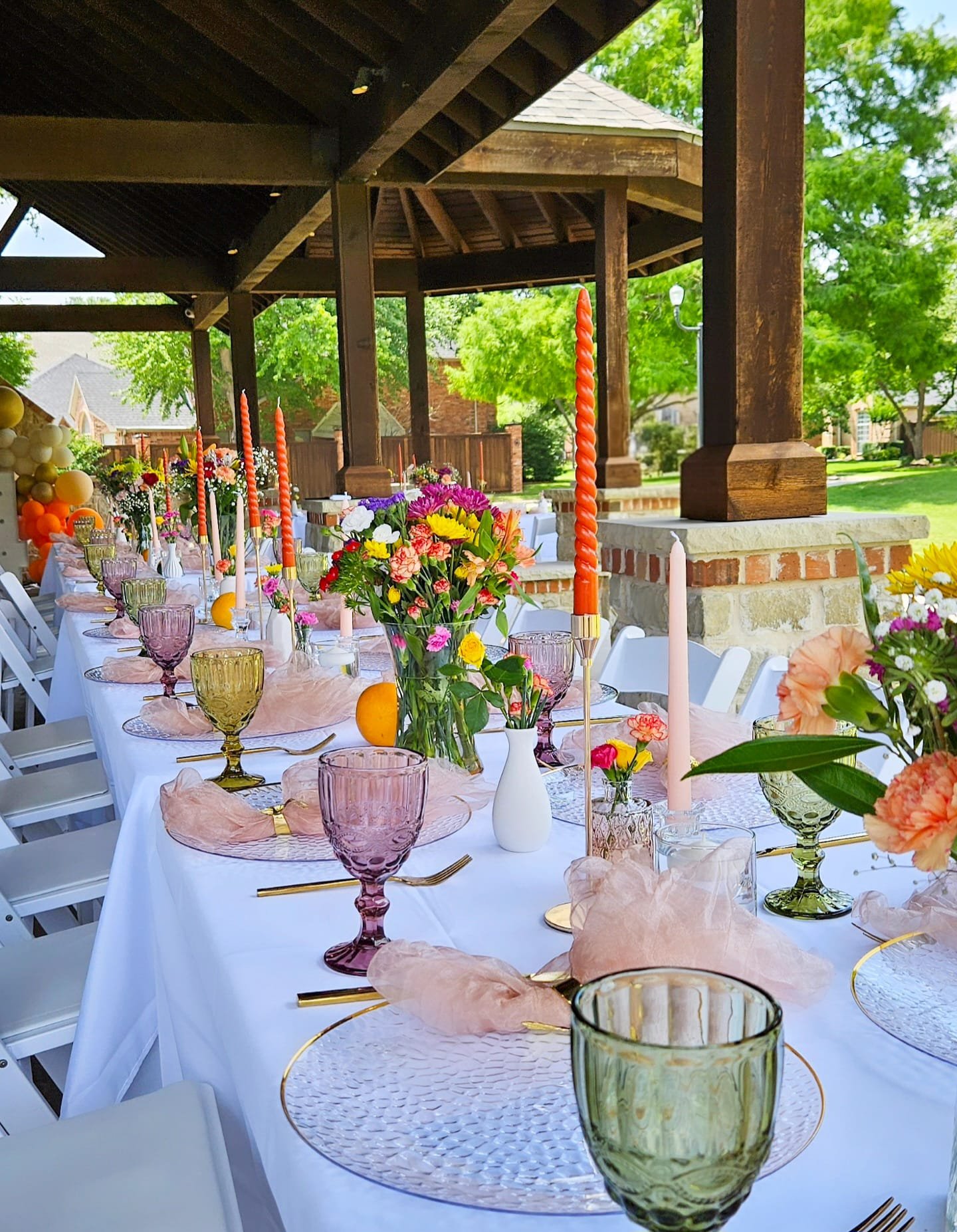 Decorated outdoor party table with pink and purple glasses, floral centerpieces, pink napkins, gold cutlery, and orange candles, under a wooden pavilion with green trees in the background.