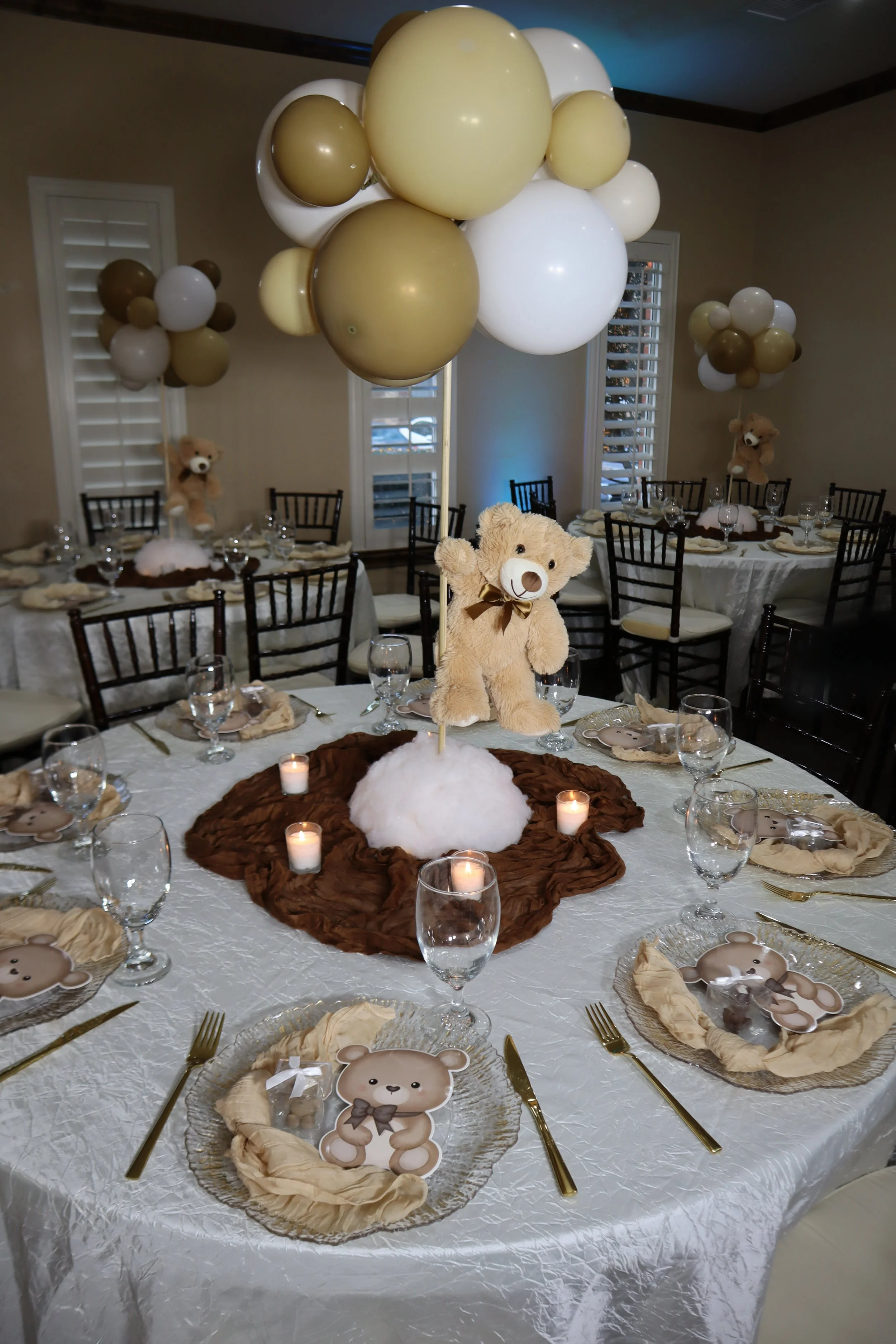 Decorated table with teddy bear themed plates, gold utensils, napkins, and small candles, with a centerpiece of plush teddy bear and balloons in white, gold, and brown, in a banquet room.