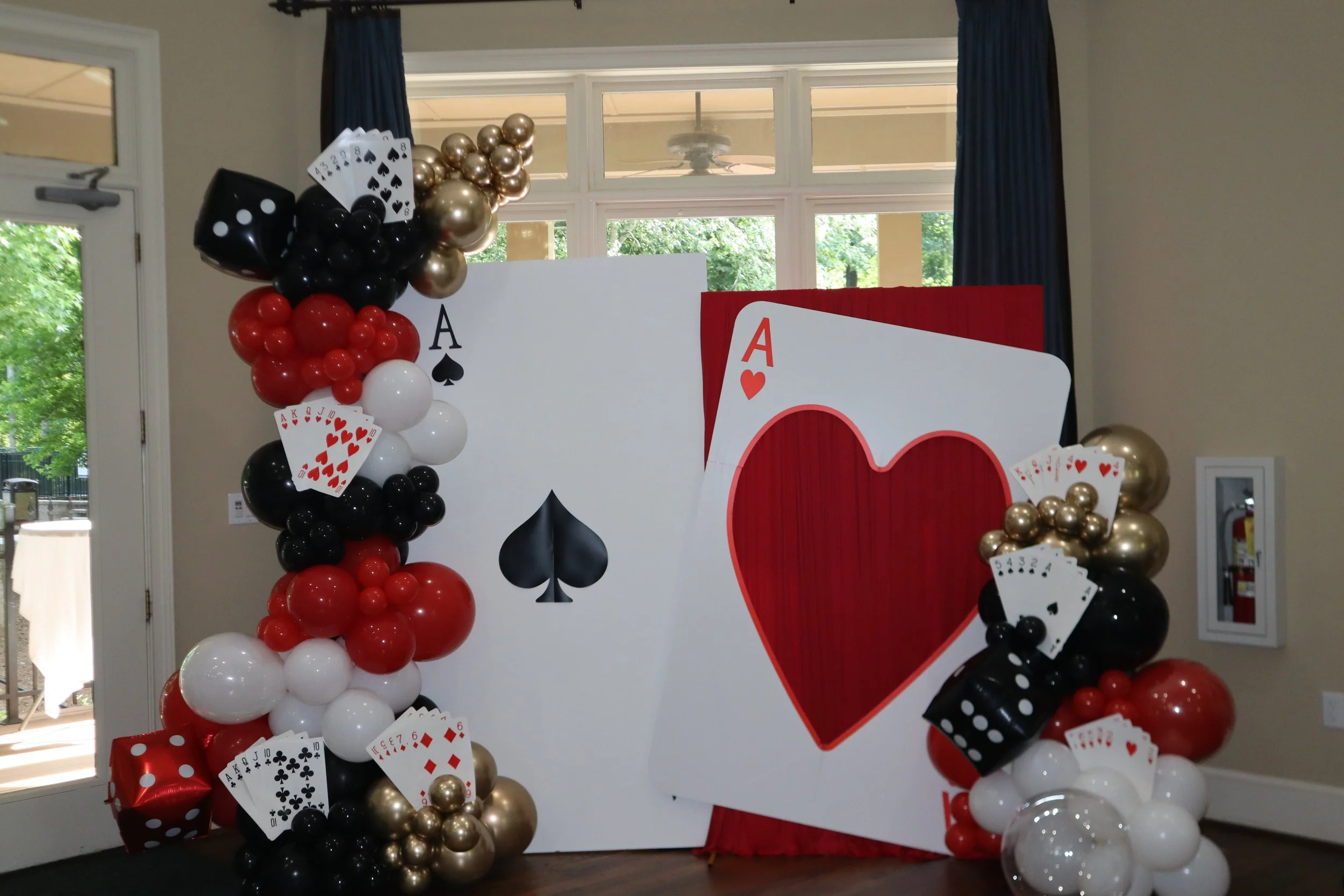Decorative setup with playing card theme, balloons in black, red, white, and gold, and large playing cards including the Ace of Spades, Ace of Hearts, and other cards, arranged around a white and red backdrop.