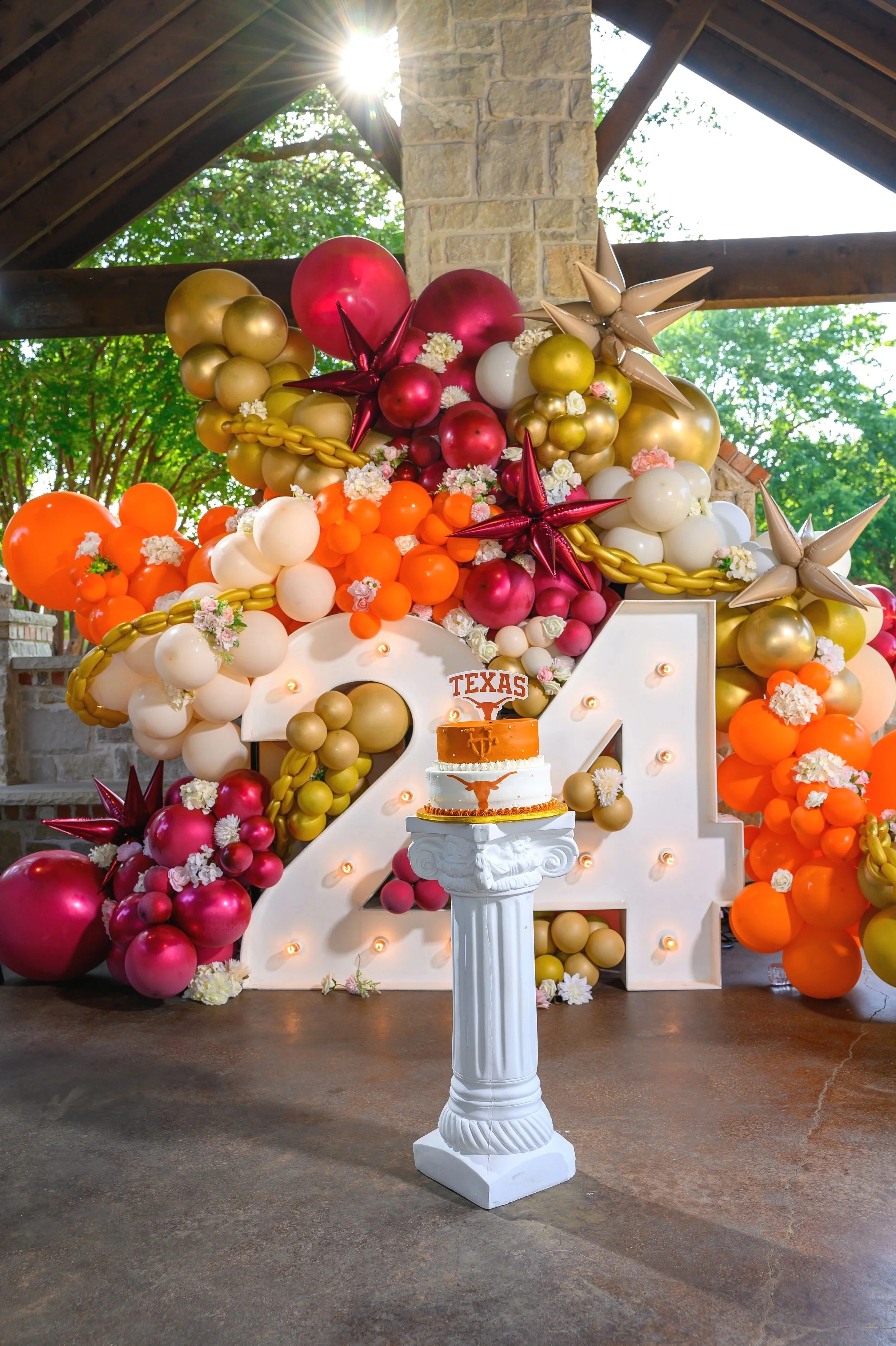 Colorful balloon arrangement with orange, white, red, and gold balloons, decorated with flowers, displayed behind a large white number 21 with lights. A cake with the Texas Longhorns logo is on a pedestal in front. The setting is outdoors with a brick and stone structure and greenery in the background.