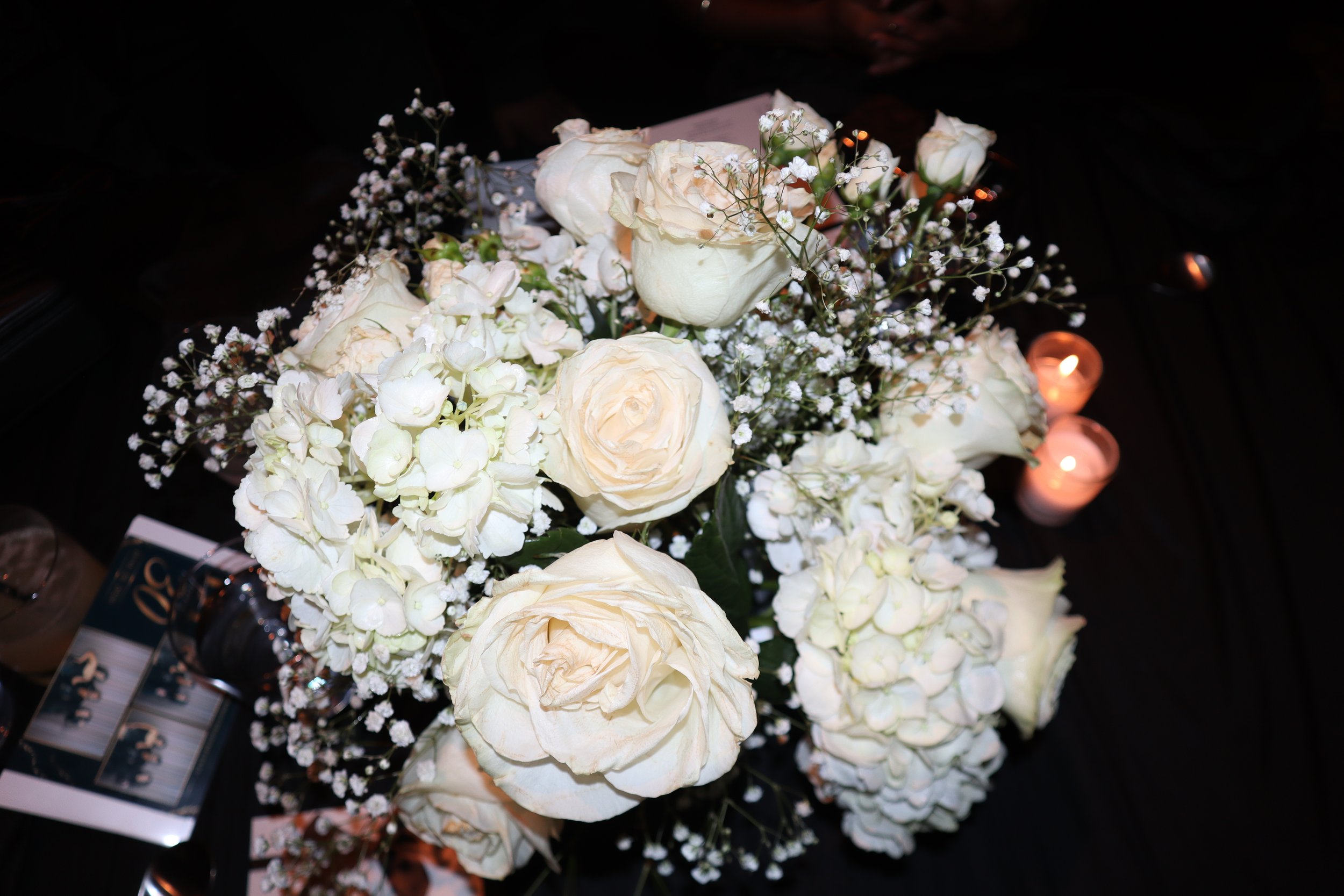 A bouquet of white roses, baby's breath, and other small white flowers with candles in small glass holders in the background.