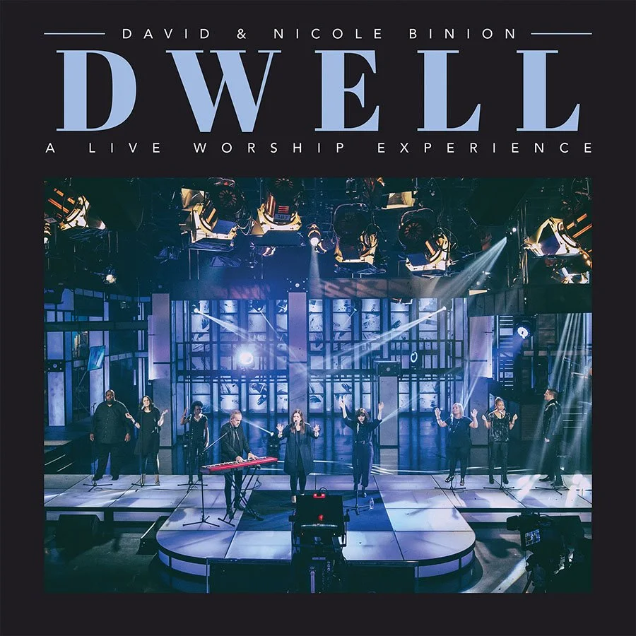 A stage with eight performers, some with microphones, and one playing a keyboard in front. The background features a large, illuminated set with multiple lights and screens. The photo is part of promotional material for the "Dwell: A Live Worship Experience" featuring David and Nicole Binion.