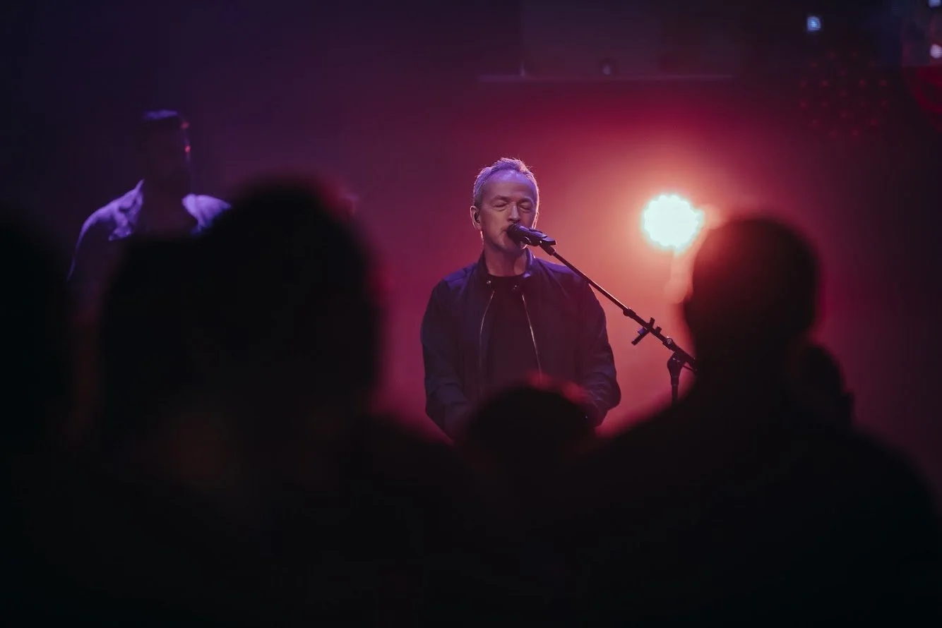 Musician with blond hair singing into a microphone on stage, illuminated by red and orange lighting, with a blurred figure of another person in the background, and an audience silhouette in the foreground.
