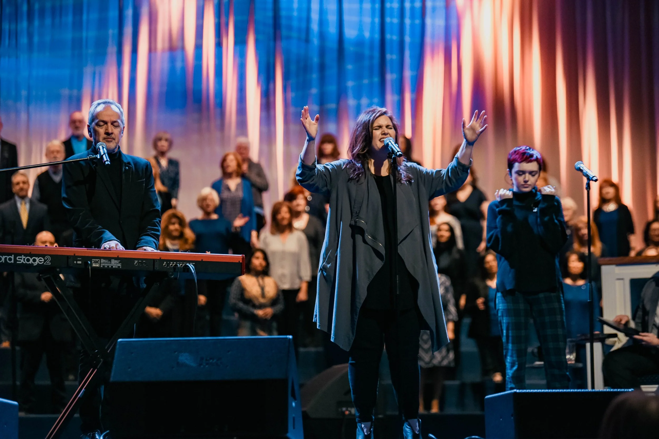 A woman singing with her eyes closed, standing with her hands raised, in a stage setting with a choir background and colorful lighting, accompanied by a keyboardist on the side.