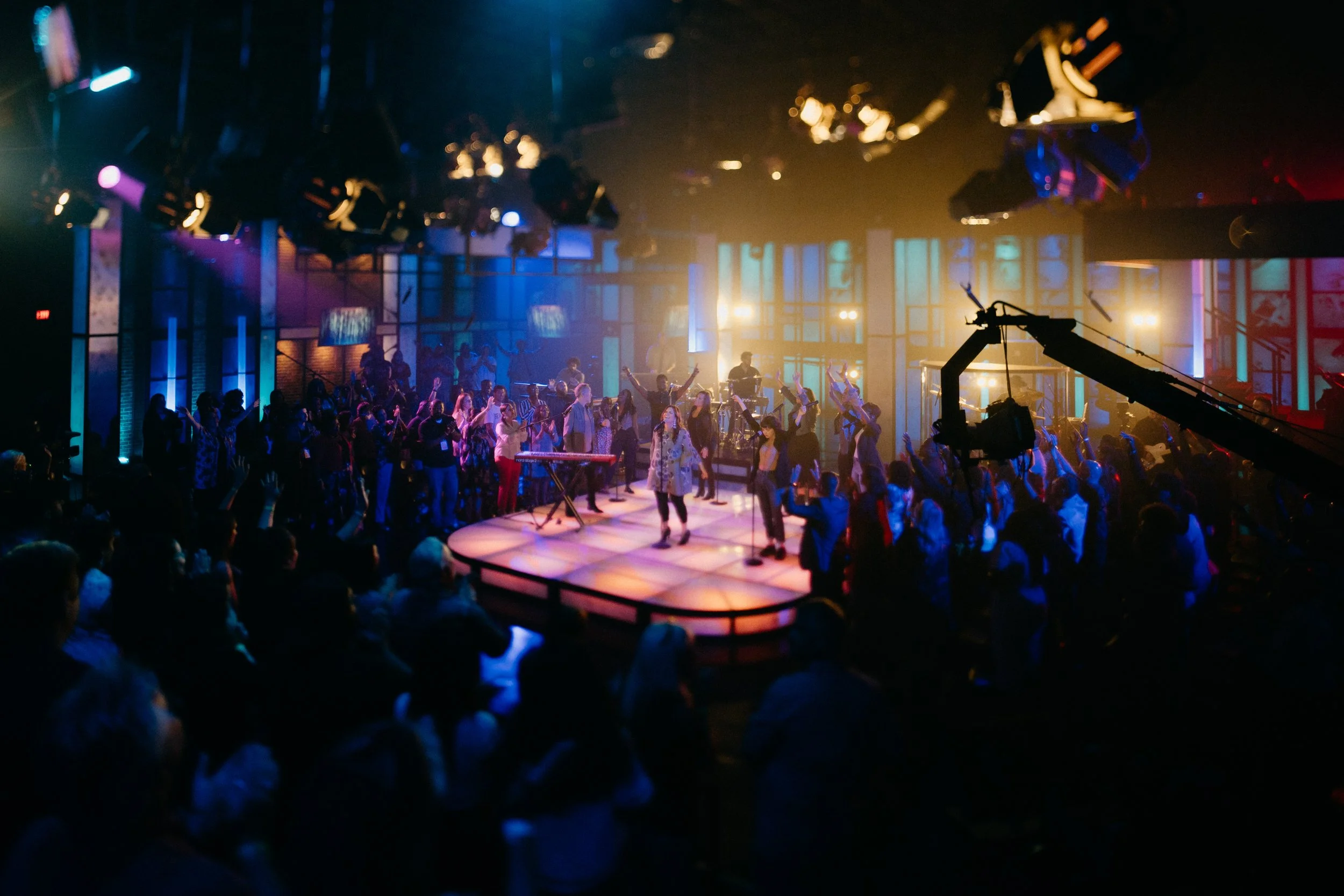 Concert or nightclub scene with people dancing and socializing under colorful stage lighting, with musical instruments and equipment on stage.