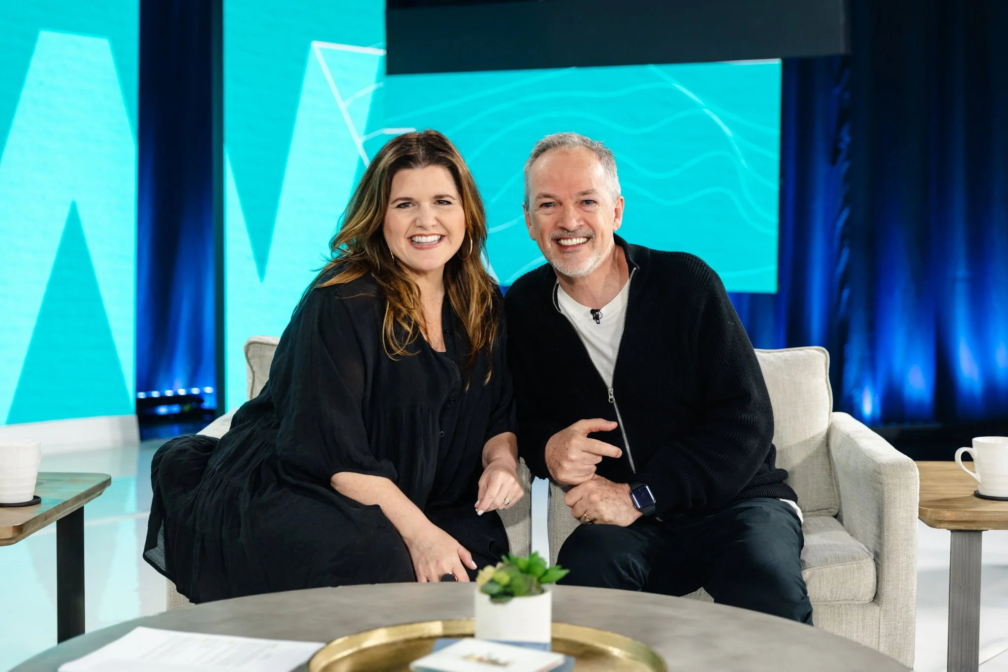 A smiling woman with long brown hair and a man with short gray hair and a beard sitting closely together on a light-colored couch, with a blue abstract background on a large screen behind them, in a modern room with a coffee table and mugs in the foreground.