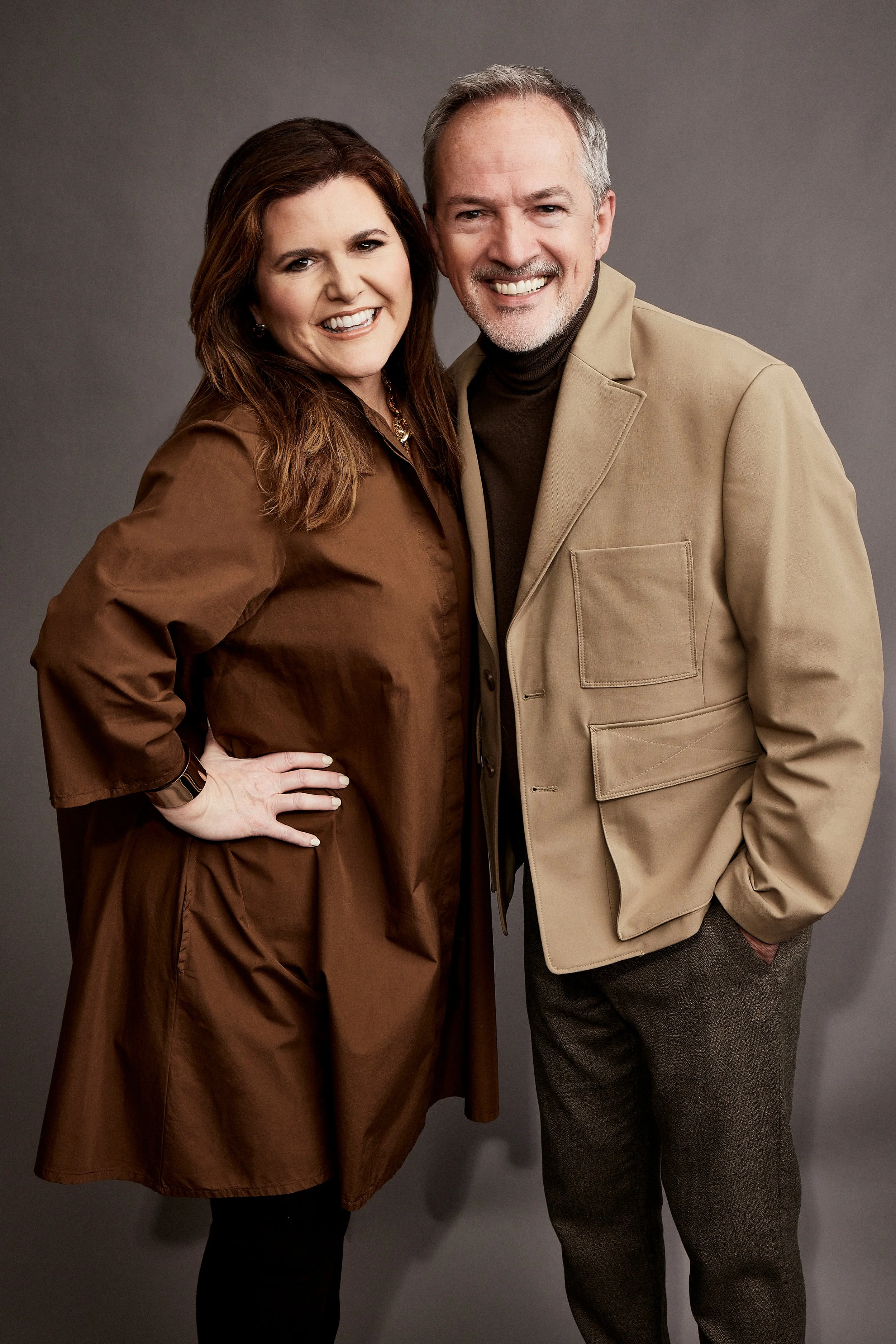 A smiling middle-aged man and woman standing close together against a gray background, dressed in professional attire.