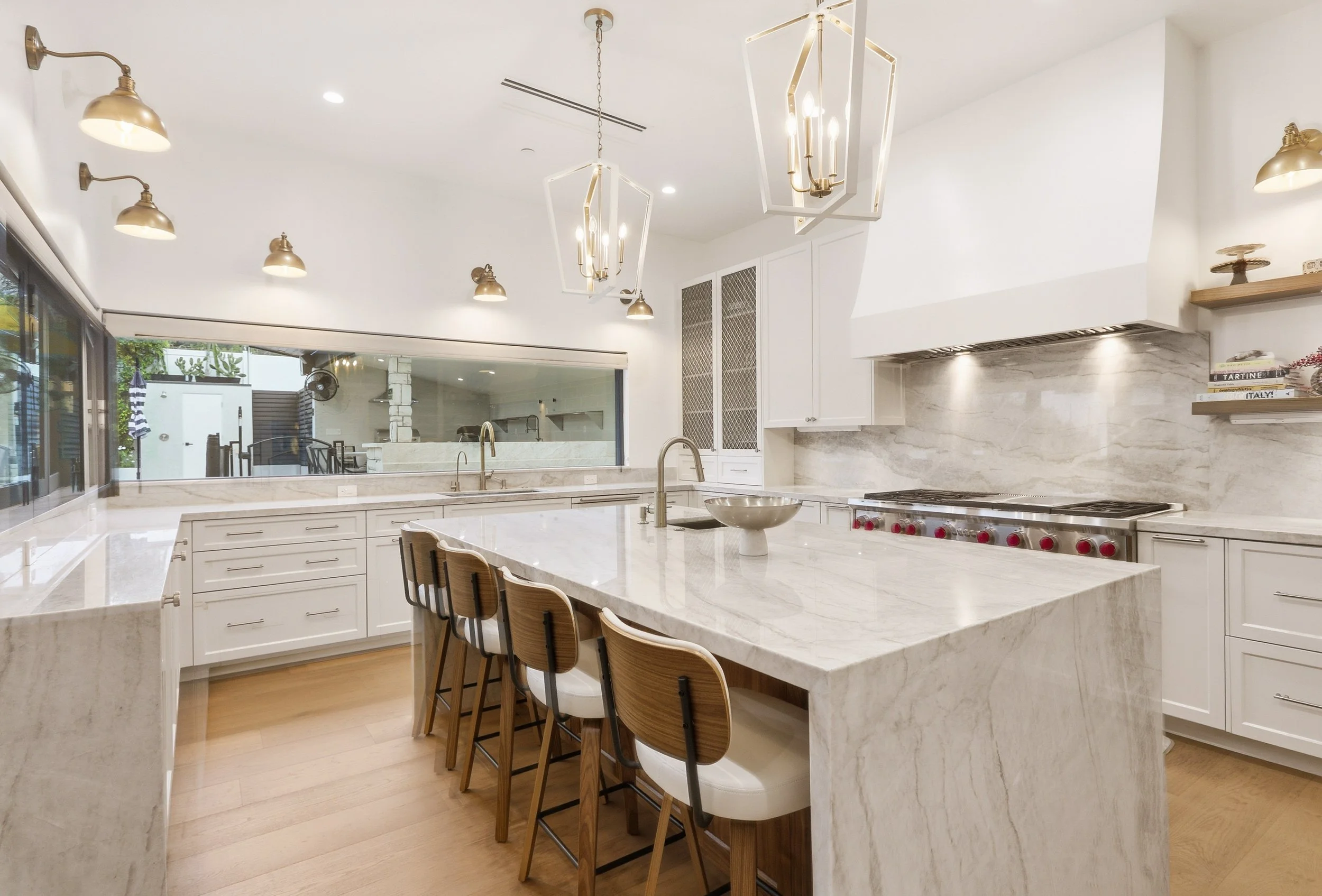 Modern kitchen with white cabinets and marble countertops, island with barstools, stainless steel appliances, pendant and wall lights, and large window showing outdoor patio with chairs and potted plants.