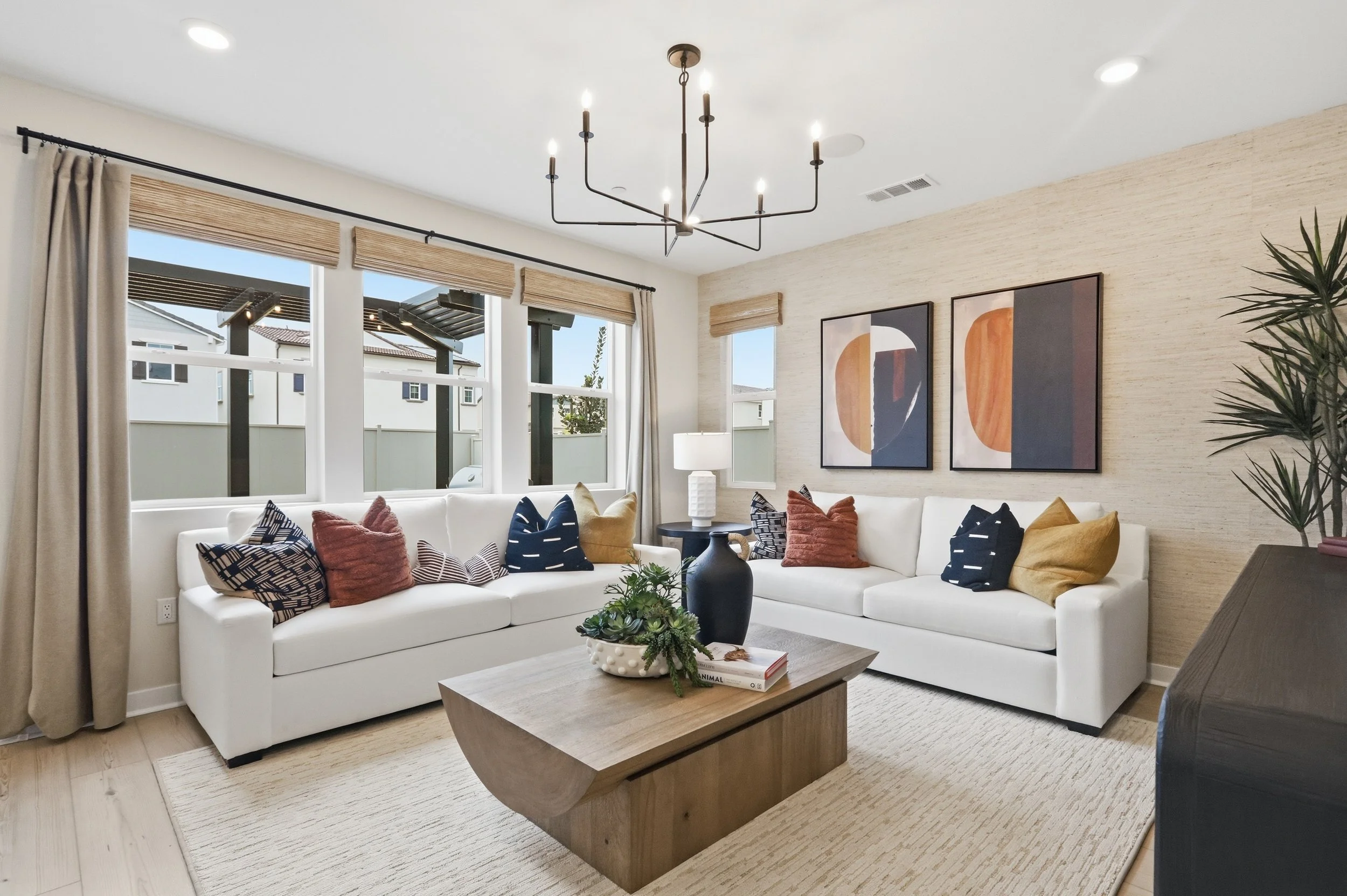 Living room with white sectional sofa decorated with assorted colorful pillows, a wooden coffee table with books and a plant, wall art, large windows with curtains, a chessboard, and a modern chandelier.