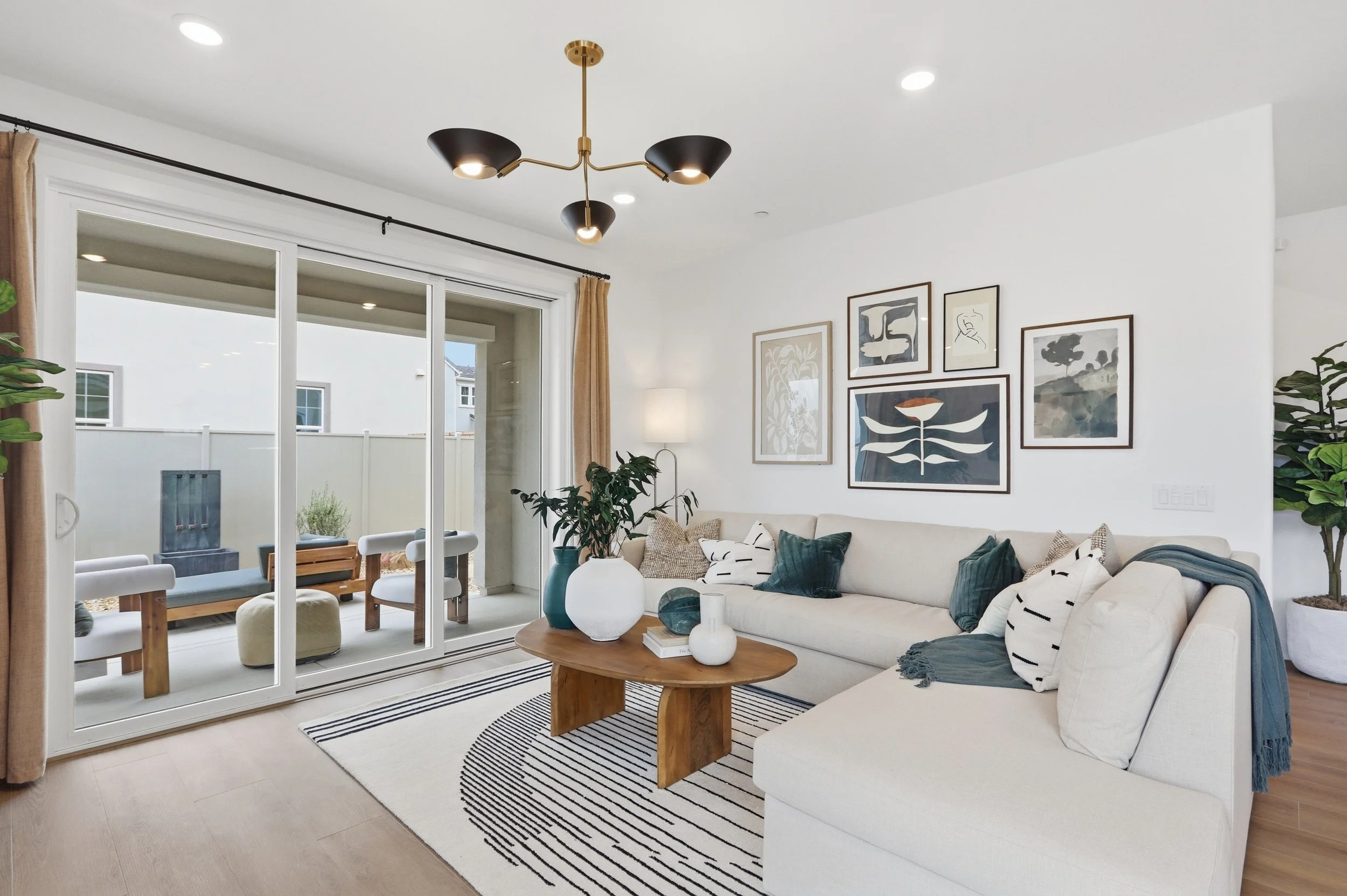 A modern living room with a white sofa decorated with black and white pillows, a wooden coffee table with vases and books, a black and white striped rug, artwork on the walls, ceiling lights, and sliding glass doors leading to an outdoor patio with w