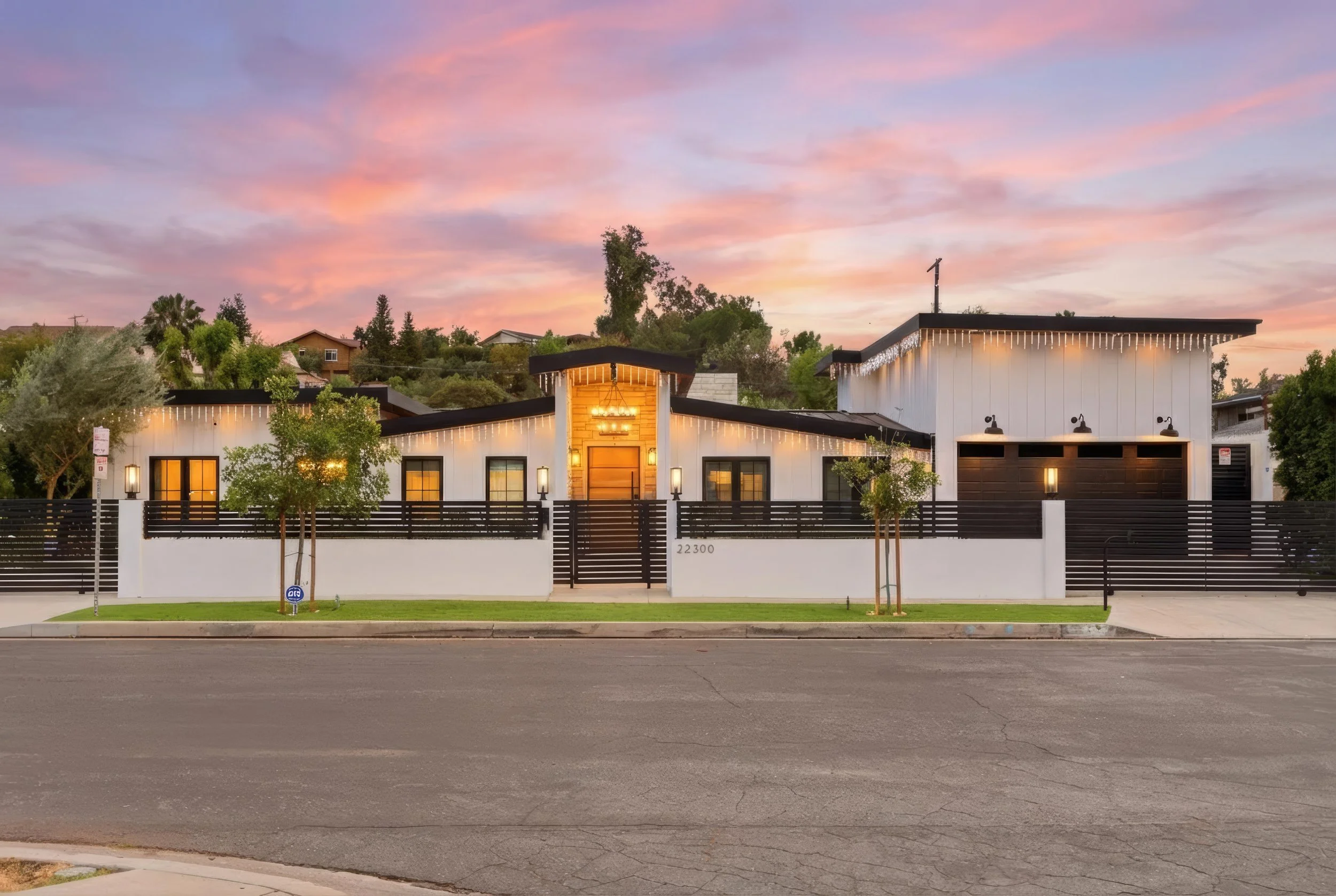 Modern white house with black accents, string lights, and a gated front yard, set against a colorful sunset sky.