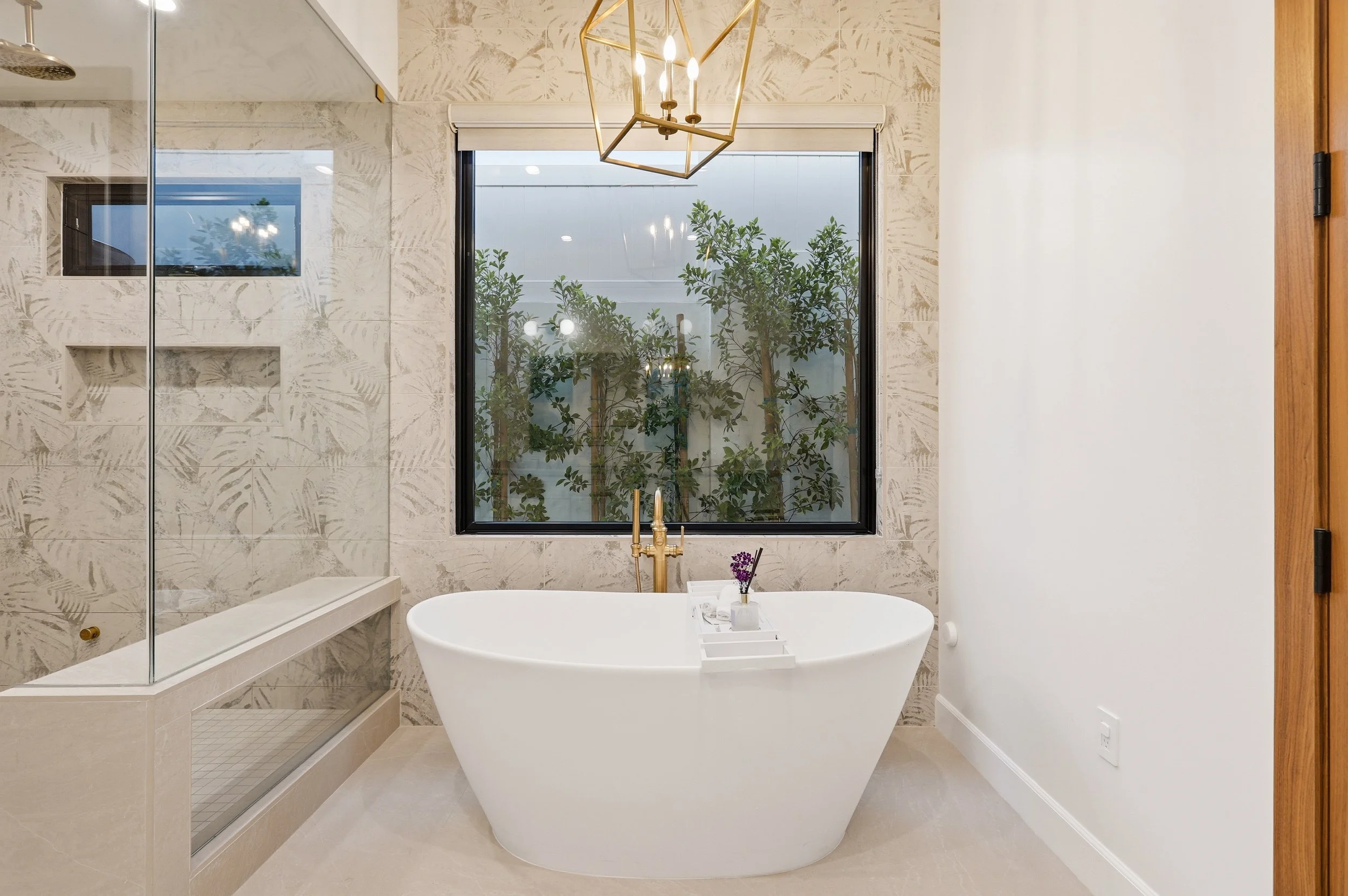 Modern bathroom with a white freestanding bathtub, large window with greenery outside, beige tiled walls with leaf patterns, a shower area with a glass door, and a contemporary gold geometric chandelier.