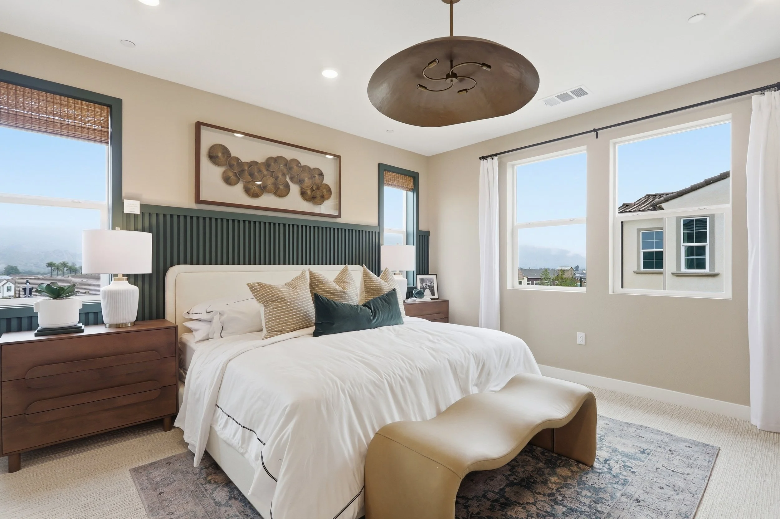 Bright bedroom with large windows, beige walls, dark green trim, white curtains, king-sized bed with white bedding, multiple pillows, wooden nightstands, lamps, wall art, and an upholstered bench at the foot of the bed.