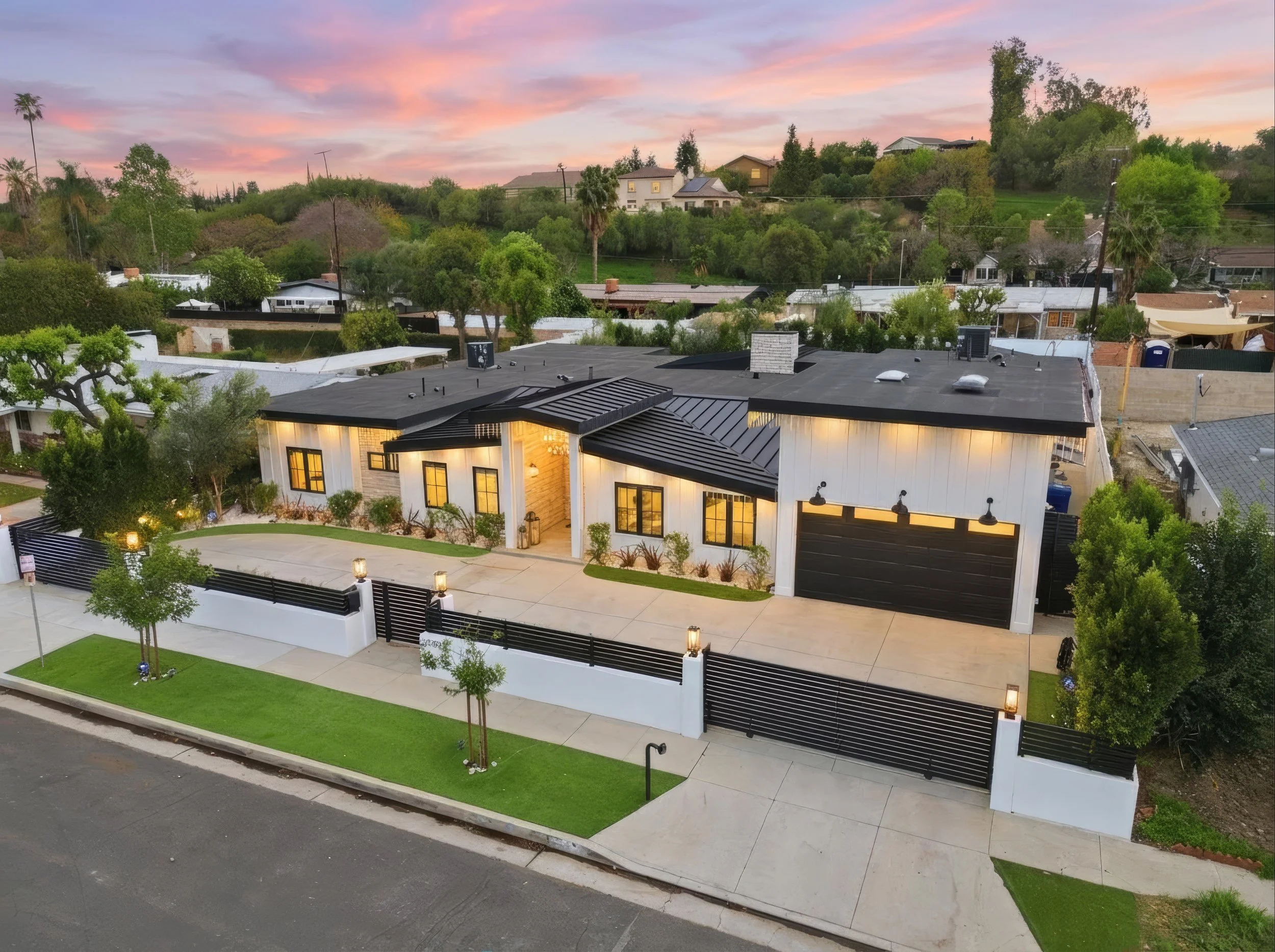 A modern house with large glass windows and a black roof in a residential neighborhood during sunset, with a green lawn and small trees in the front yard.