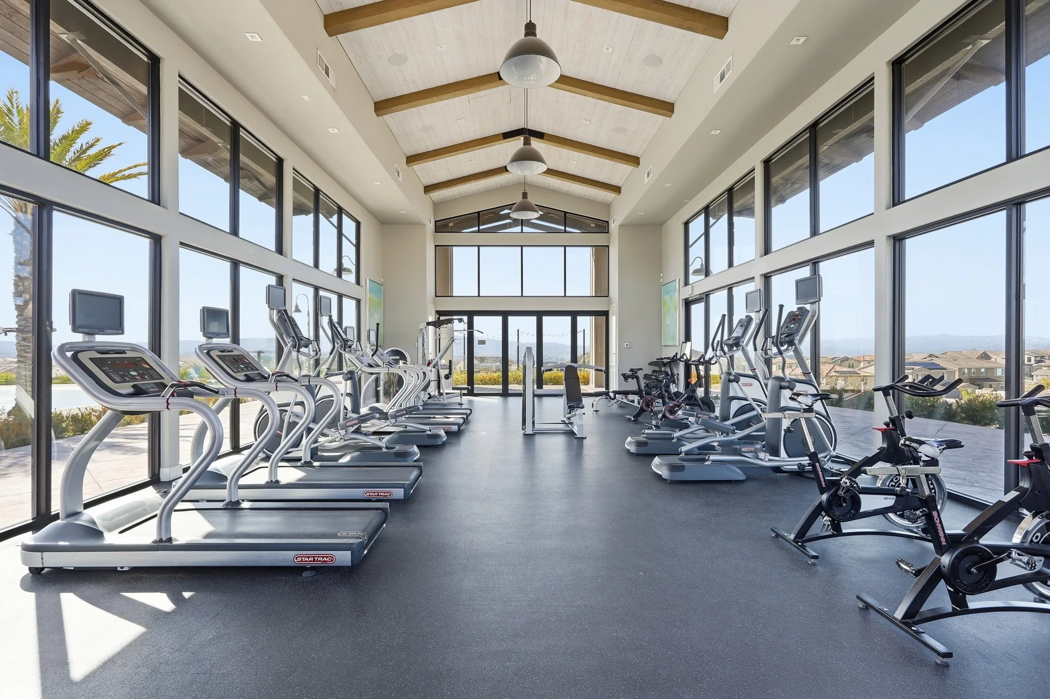 A spacious gym with large windows, equipped with treadmills, exercise bikes, and other fitness equipment, overlooking a sunny outdoor landscape.