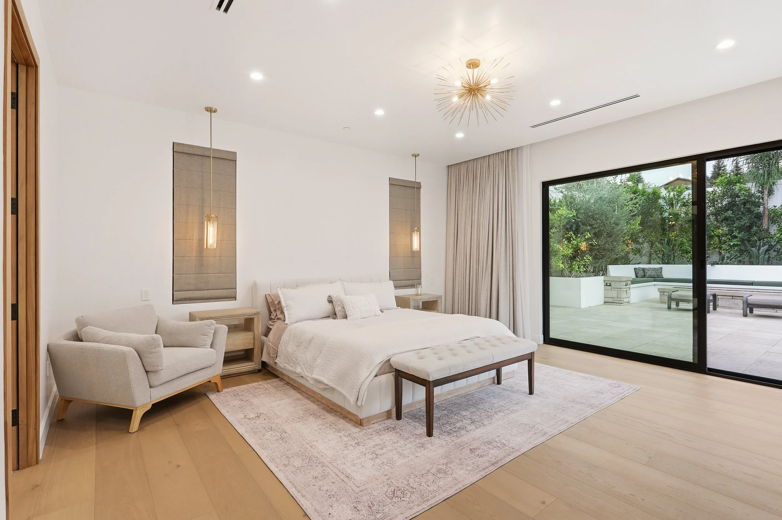 A modern bedroom with a large bed, a beige armchair, and sliding glass doors leading to an outdoor patio. The room has white walls, wooden flooring, and contemporary lighting fixtures.