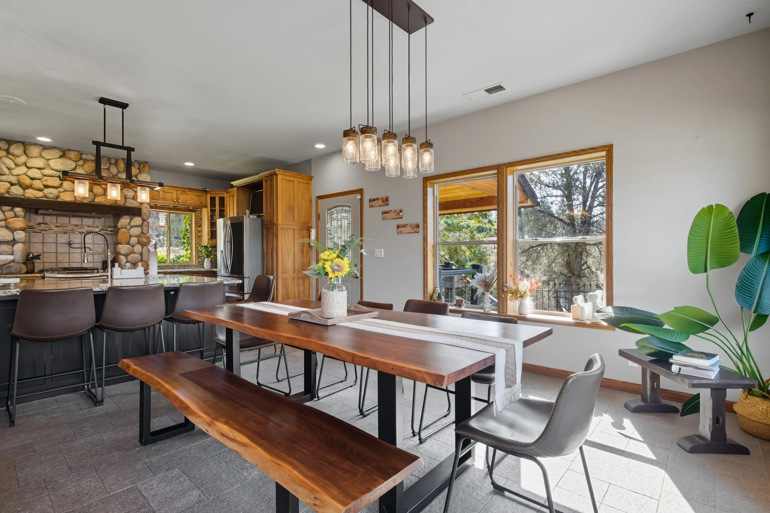 A bright kitchen and dining area with large windows bringing in natural light. It features a wooden dining table with a centerpiece of yellow flowers, modern black chairs, and a rustic stone fireplace. There are stylish pendant lights hanging above t