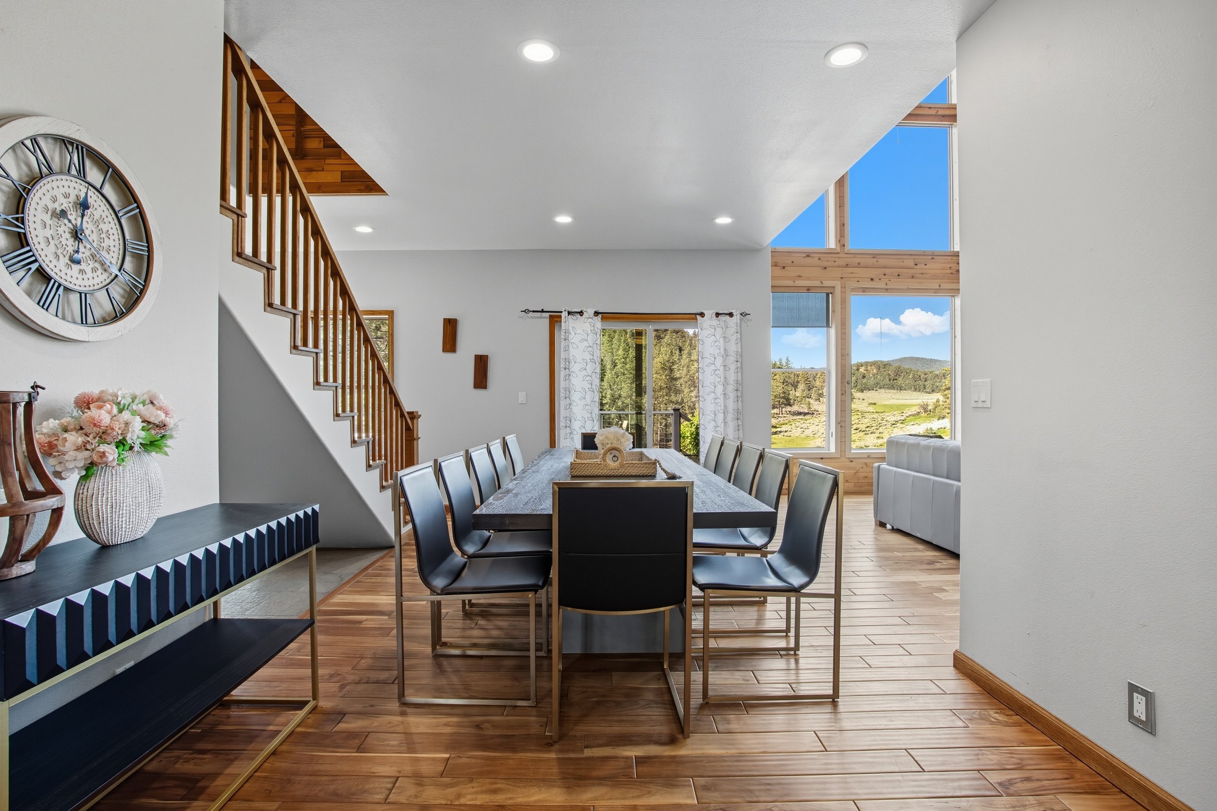 Modern open-concept dining area with large windows, wooden flooring, and a mix of contemporary and rustic furniture, including a dining table with chairs and a sideboard with a flower vase.